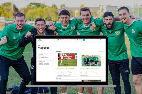 F&uuml;nf l&auml;chelnde M&auml;nner in passenden gr&uuml;nen Fu&szlig;balltrikots posieren gemeinsam drau&szlig;en auf einem Feld. Im Vordergrund zeigt ein Computermonitor eine Website mit Artikeln und Bildern zum Thema Fu&szlig;ball an.