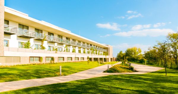 Das Akademia Sporthotel ist ein modernes Hotel mit Balkonen, das direkt an einem gepflasterten Fußweg und einer üppig grünen Rasenfläche liegt. Es ist von Bäumen umgeben und befindet sich unter einem strahlend blauen Himmel mit vereinzelten Wolken. Hier k