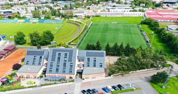 Luftaufnahme des Hotels Benica***, eines modernen Gebäudes mit Sonnenkollektoren auf dem Dach, das neben zwei grünen Kunstrasen-Fußballplätzen liegt. Im Hintergrund sind Bäume, geparkte Autos und eine Stadtlandschaft zu sehen.