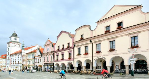Radfahrer fahren über einen historischen Stadtplatz in einer europäischen Stadt, umgeben von pastellfarbenen Renaissancegebäuden, gewölbten Arkaden und dem hohen Uhrturm des Hotels „Zlata Hvezda“, der im Hintergrund unter einem bewölkten Himmel stolz empo
