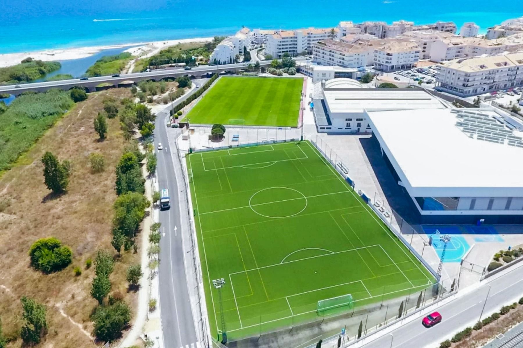 Luftaufnahme eines Sportkomplexes an der Küste in der Nähe des Hotels SH Villa Gadea*, mit grünen Fußballplätzen, die sich ideal für Trainingslager in Altea und Fußballmannschaften eignen, umrahmt von Gebäuden, einer Straße, Bäumen und dem blauen Meer.