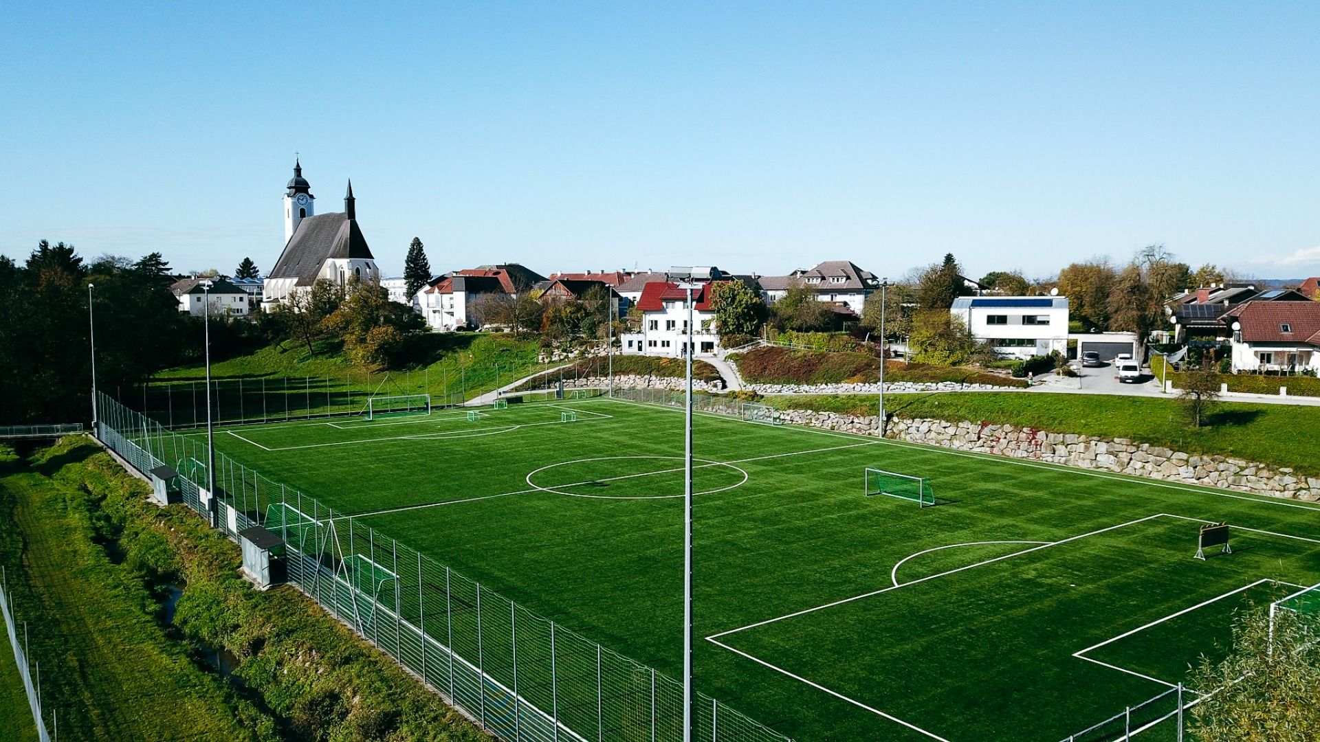 Luftaufnahme des grünen Kunstrasenplatzes in Neuhofen an der Ybbs, in der Nähe des Perbersdorfer Heurigen - Hotels. Umgeben von einem Zaun mit kleinen Toren, Häusern und einem großen Kirchturm unter einem klaren blauen Himmel.
