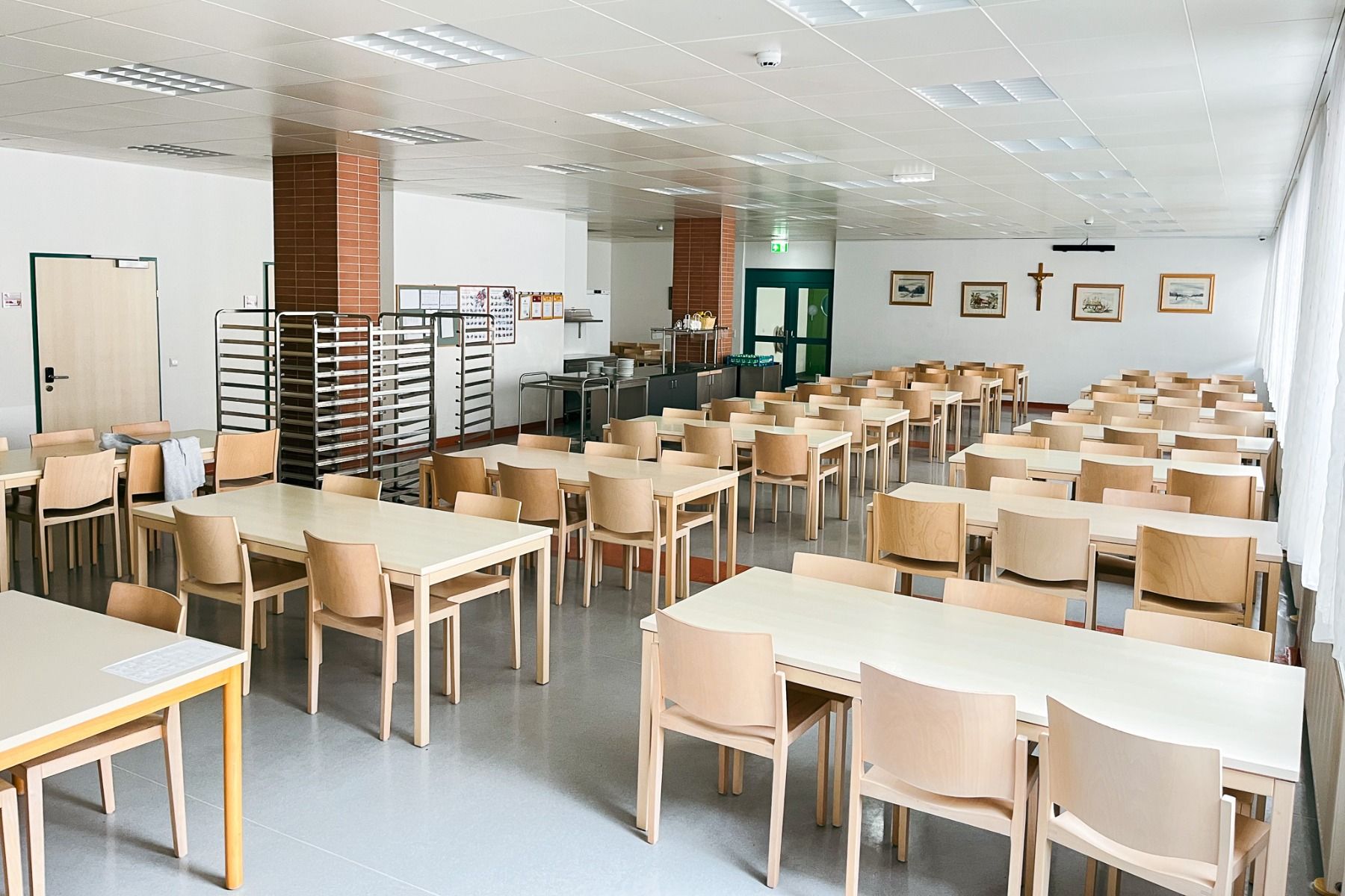 Das Gästehaus Burgenland Güssing verfügt über eine helle Cafeteria mit hellen Holztischen und -stühlen, die ordentlich angeordnet sind, Metallregale an den Wänden, gerahmte Bilder, ein Kruzifix und große Fenster, die den Raum mit natürlichem Licht füllen.