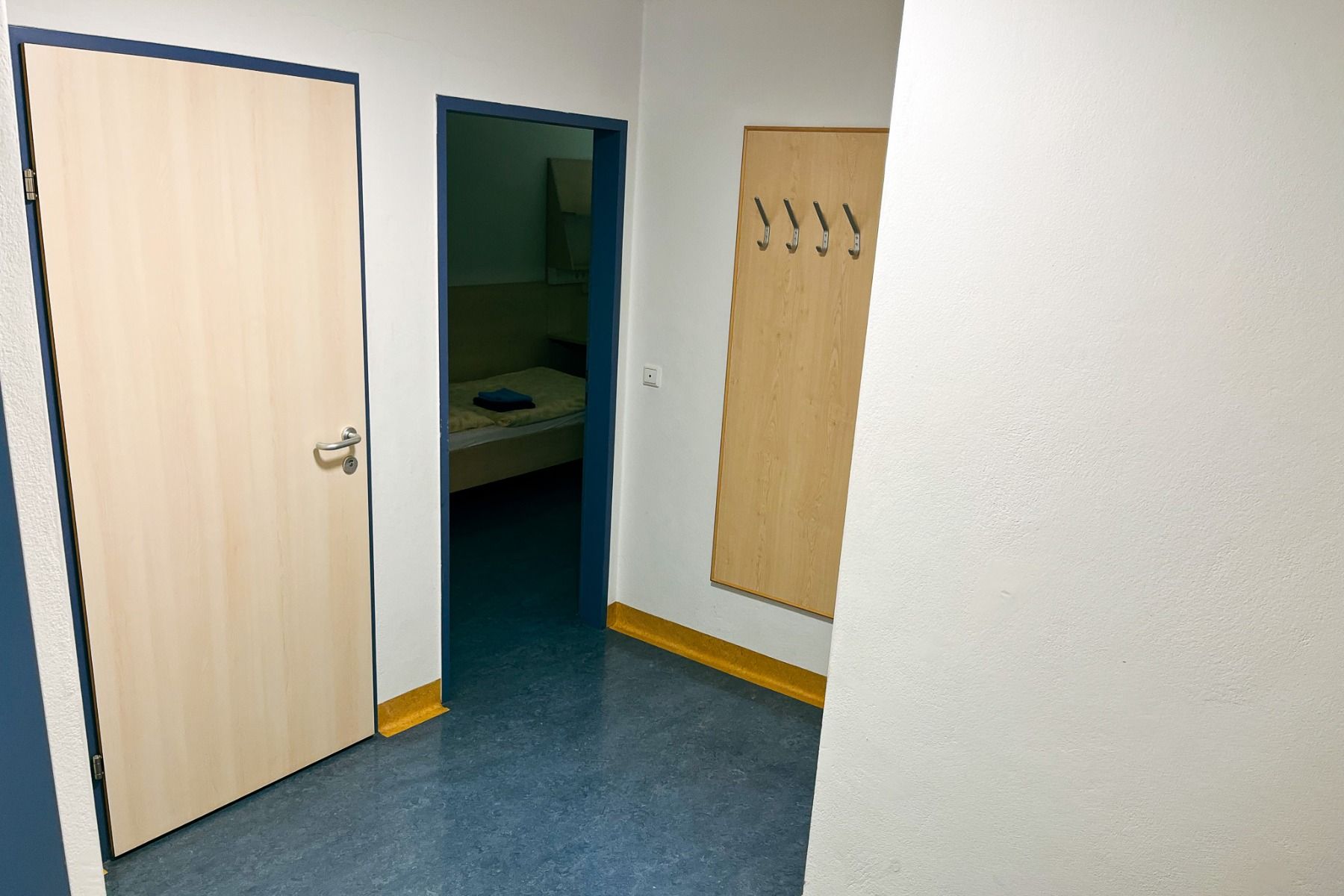 Im Gästehaus Burgenland Güssing gibt es einen Flur mit blauem Boden, eine helle Holztür auf der linken Seite, eine offene Tür, in der ein Bett zu sehen ist, und eine Wandgarderobe mit fünf Haken auf der rechten Seite.