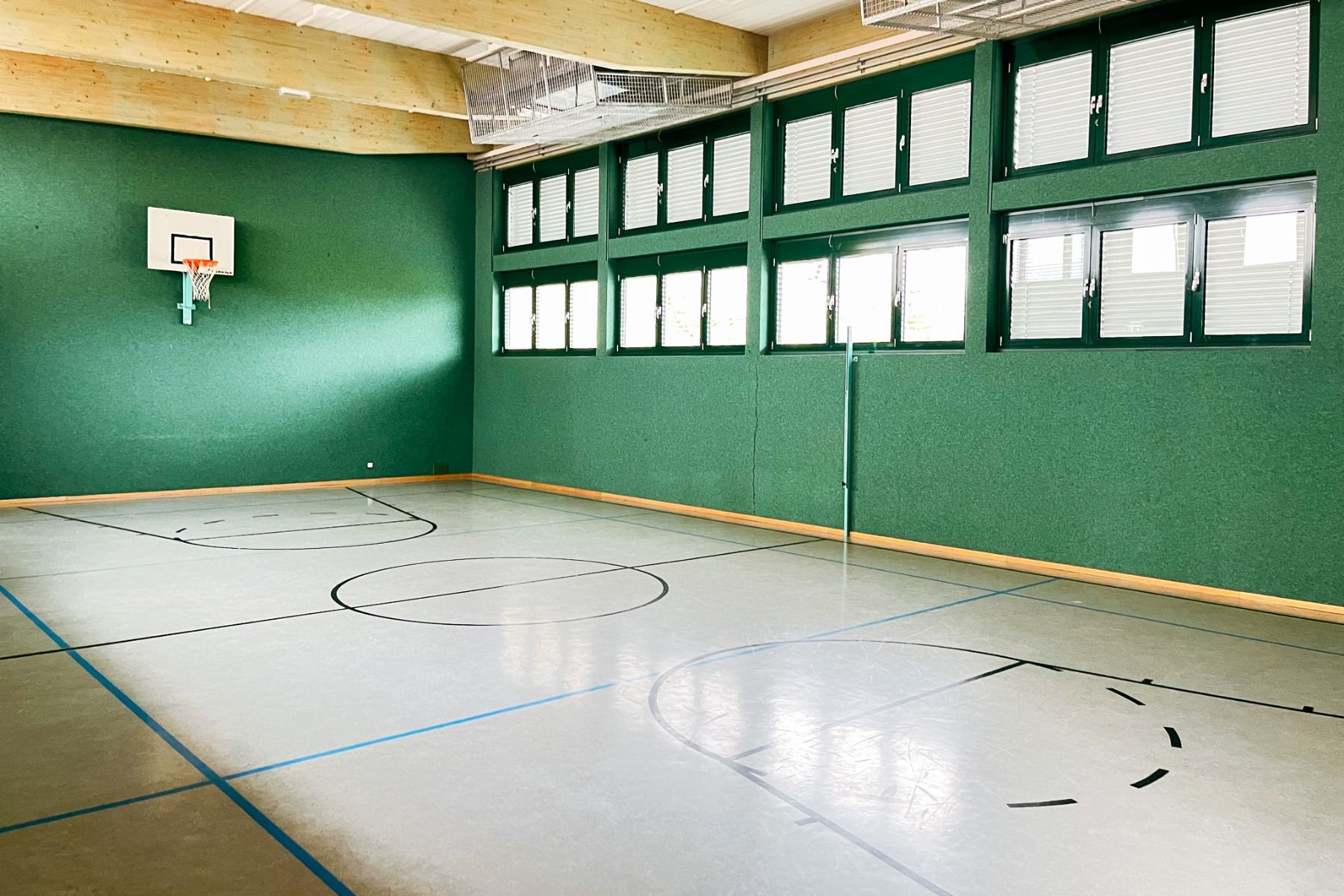 Das Gästehaus Burgenland Güssing verfügt über eine Turnhalle mit einem Basketballkorb, grünen Wänden, hohen Fenstern mit Jalousien und einem polierten Boden, der für den Sport gekennzeichnet ist. Durch die großen Fenster strömt das Sonnenlicht herein.