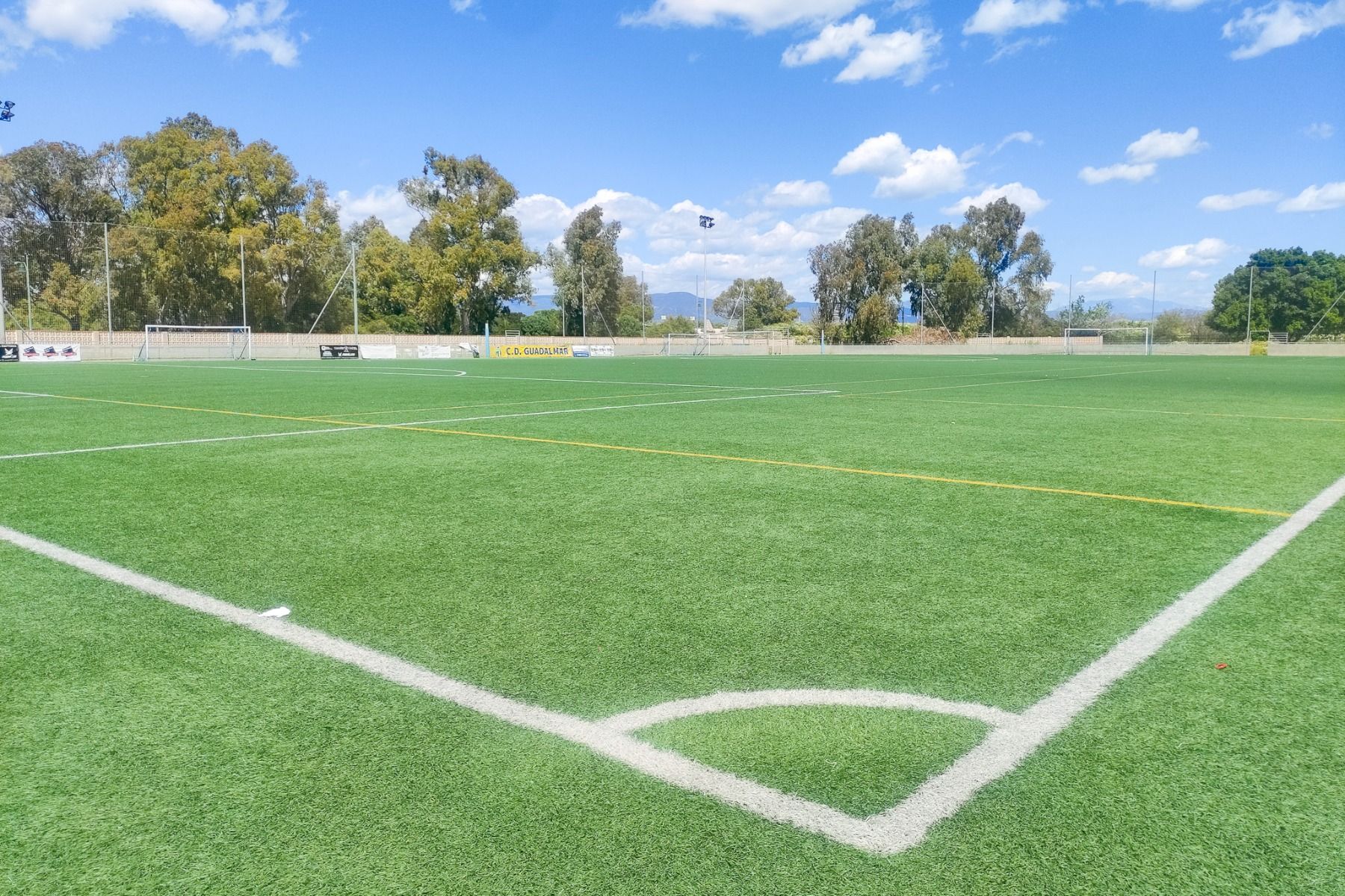 Ein klarer, sonniger Tag auf einem Fußballplatz mit grünem Kunstrasen in der Nähe des Hotels Sol Guadalmar, weiße und gelbe Markierungen, Bäume im Hintergrund und ein blauer Himmel mit vereinzelten Wolken.