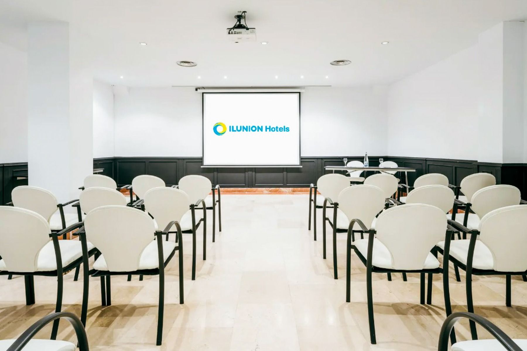 Et konferanserom på Hotel ILUNION Bel Art Barcelona har rader med hvite stoler foran en stor skjerm med hotellets logo, minimalistisk innredning og svarte paneler - perfekt for arrangementer om fotballtrening eller fotballkultur i Barcelona.