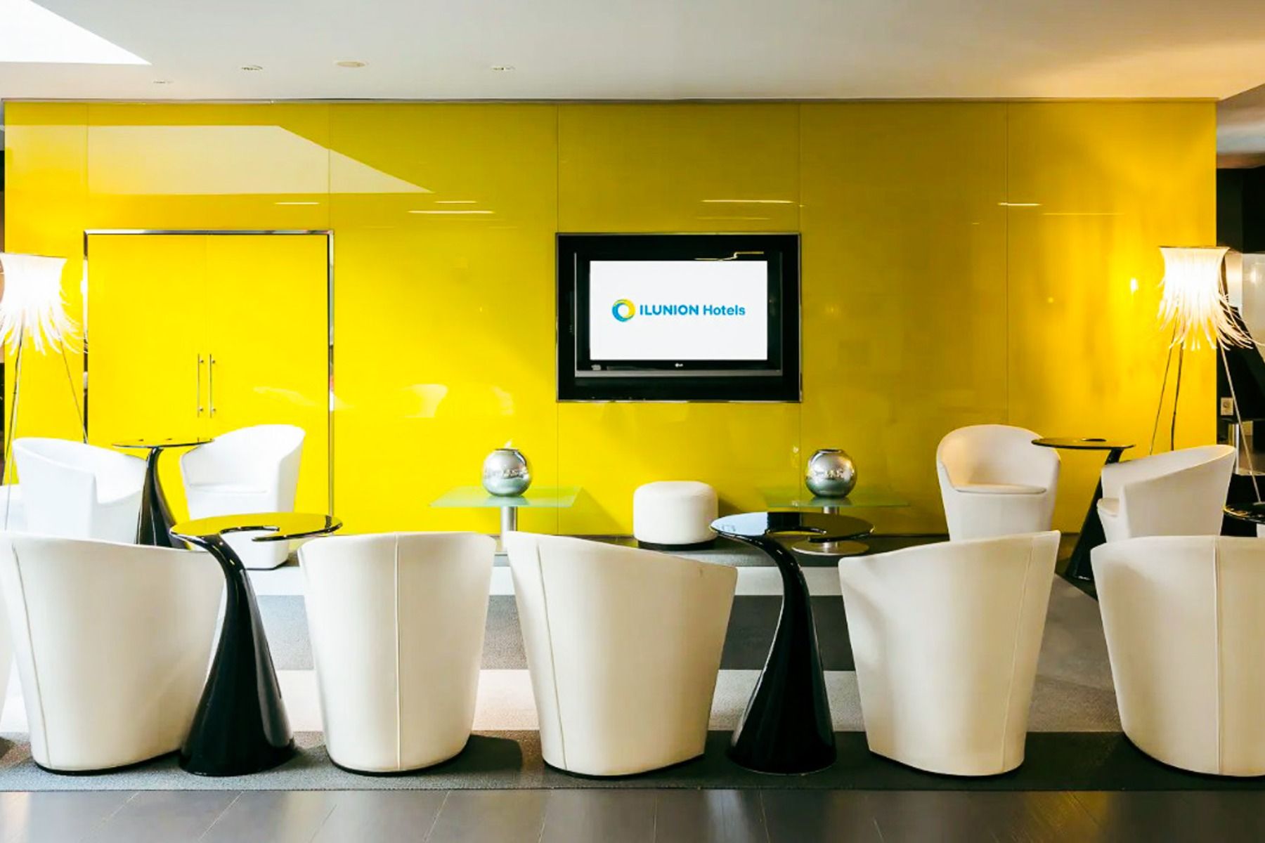 Den moderne lobbyen på Hotel ILUNION Bel Art Barcelona har hvite lenestoler, elegante svarte bord, glassinnredning og en levende gul vegg med en TV som viser ILUNION Hotels - noe som gjenspeiler Barcelonas energiske fotballkultur. To stilige lamper fullfø