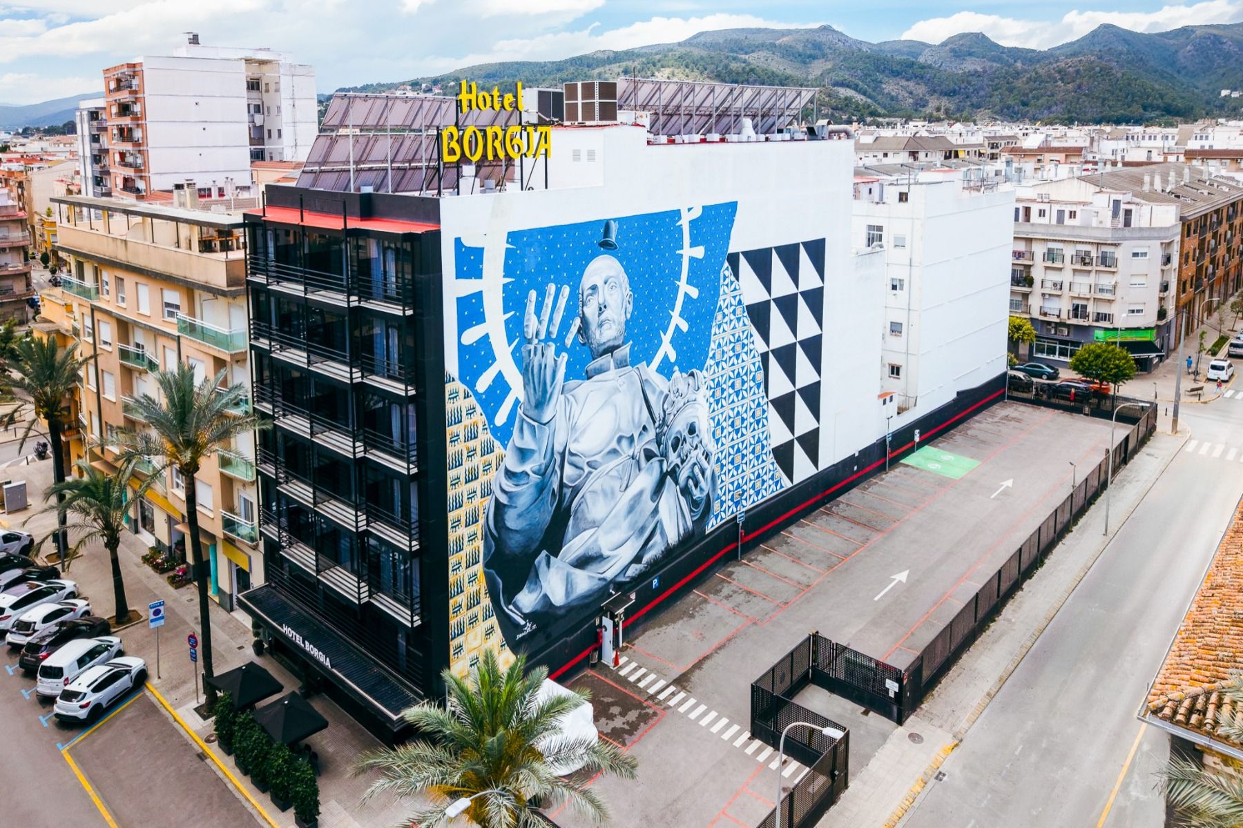 Ein großes Wandgemälde einer futuristischen Figur ziert die Fassade des Hotels Borgia *, das auf einen größtenteils leeren Parkplatz blickt, inmitten einer Stadtlandschaft, die von Hügeln und Gebäuden eingerahmt wird. Das Hotel Borgia *, das dafür bekannt