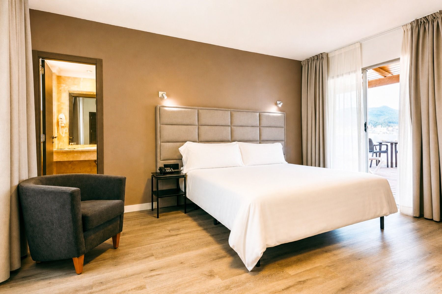 Modernes Hotelzimmer im Hotel Borgia *** mit einem Doppelbett mit grauem Kopfteil, Nachttischen, einem grauen Sessel und Holzboden. Das eigene Badezimmer ist durch eine offene Tür zu sehen. Große Fenster mit Vorhängen führen auf einen Balkon mit Blick auf