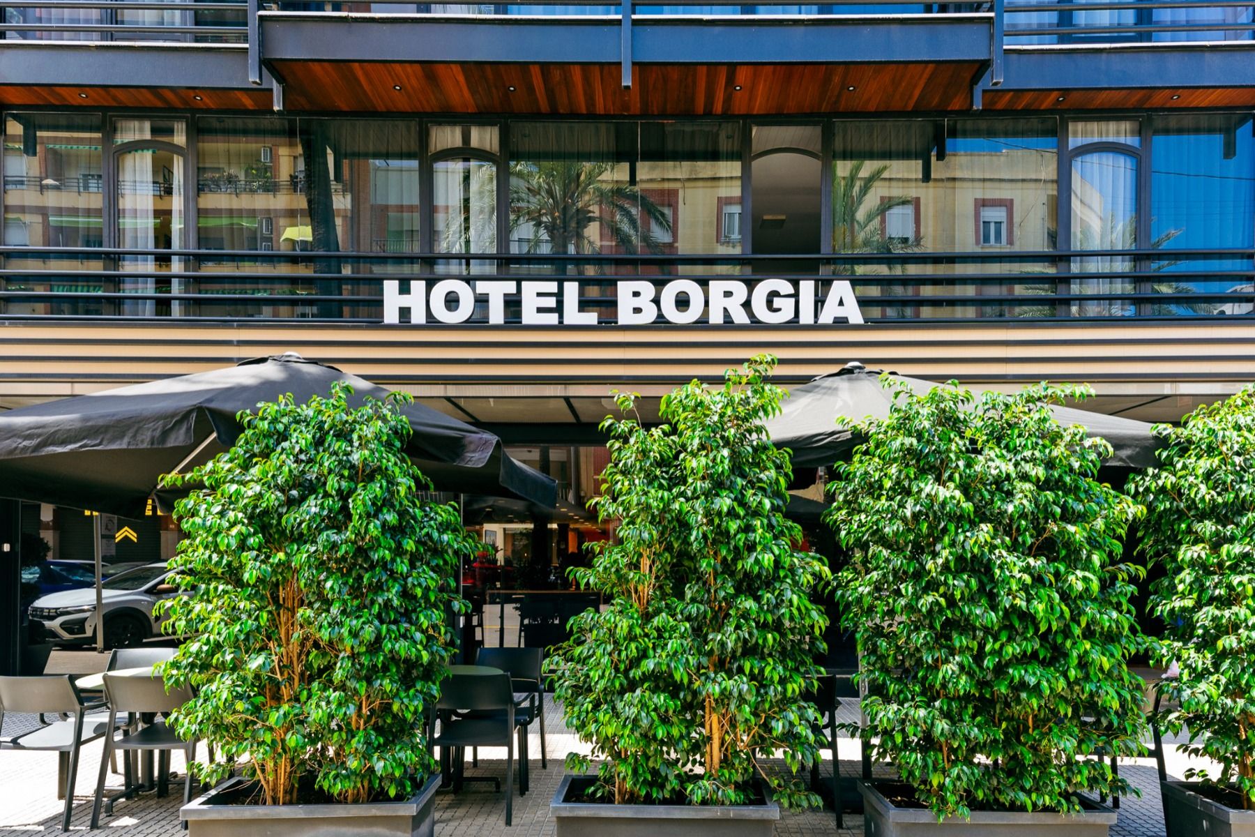 Das Hotel Borgia * besticht durch eine einladende Außenansicht mit großzügigen Glasfenstern, Sitzgelegenheiten im Freien, die von eleganten schwarzen Sonnenschirmen beschattet werden, sowie hohen Grünpflanzen in Pflanzkübeln, die die Gäste am Eingang will