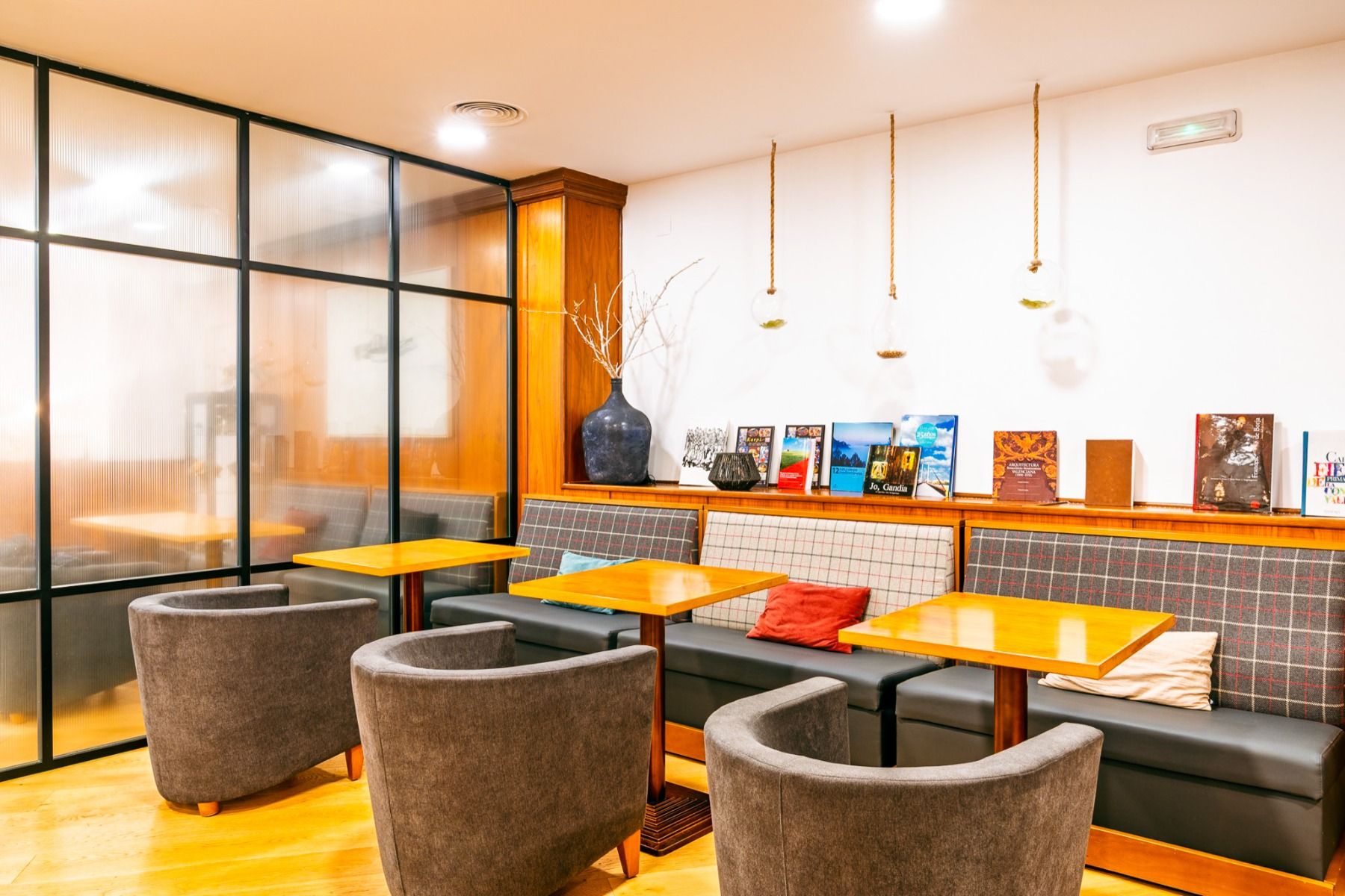 Ein gemütliches Café mit Holztischen, grauen gepolsterten Stühlen und einer karierten Bank schafft eine einladende Atmosphäre. Ausgestellte Bücher und eine Glastrennwand verleihen dem Raum zusätzlichen Charme, ergänzt durch warme Beleuchtung und dekorativ