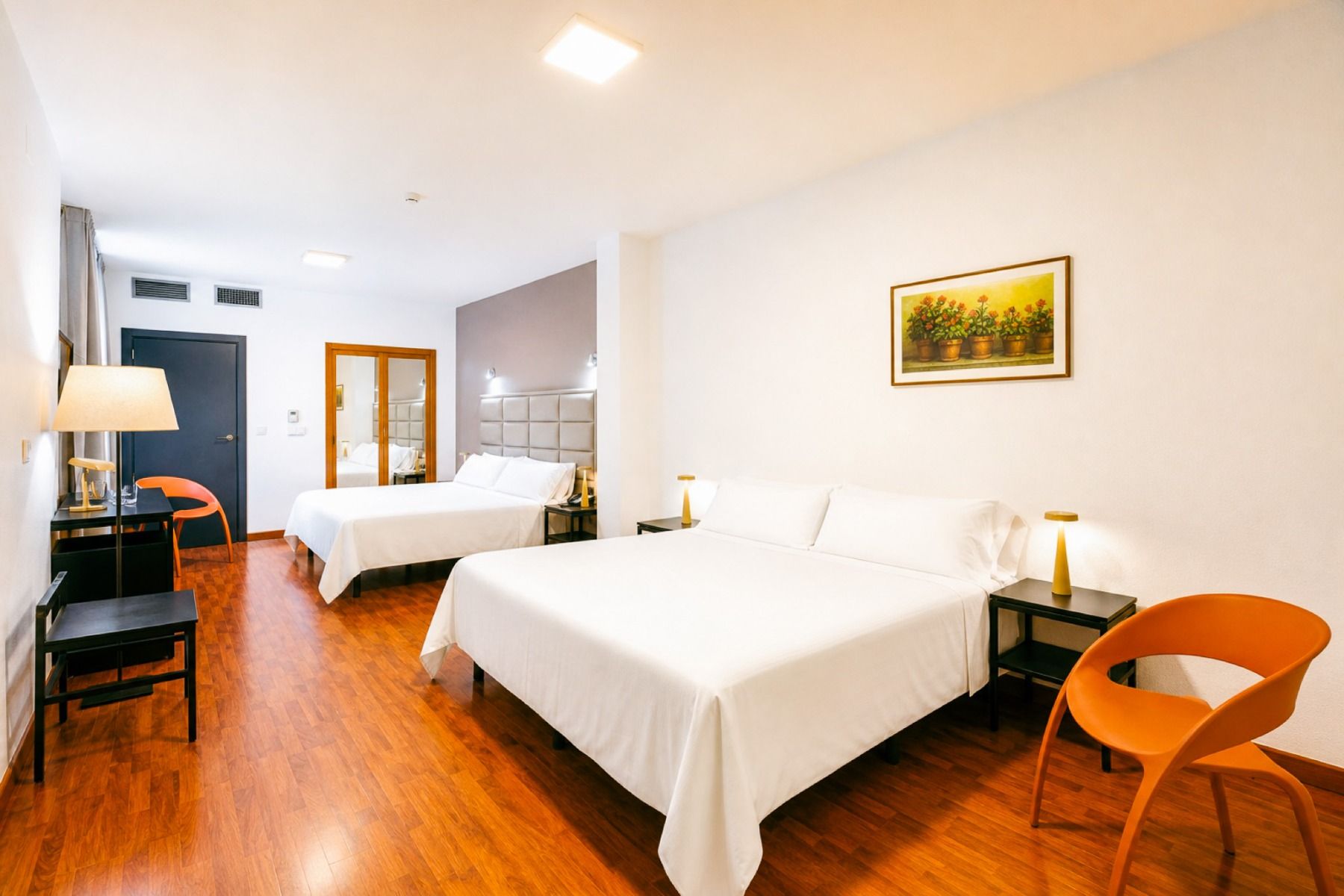 Ein helles und gepflegtes Zimmer im Hotel Borgia *** mit zwei Doppelbetten, Holzboden, zwei orangefarbenen Stühlen, einem Schreibtisch mit Lampe und einem Blumenbild an der Wand – die ideale Wahl für Fußballmannschaften, die während ihres Trainingslagers