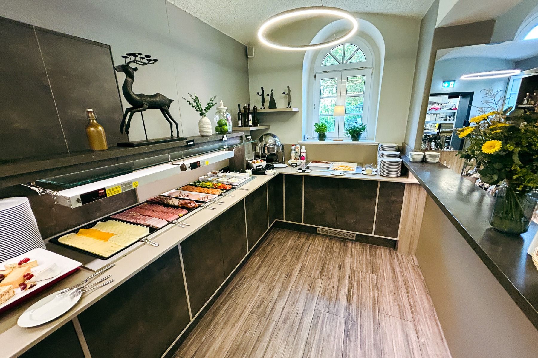 Ein Buffet mit einer Auswahl an Aufschnitt, Käse, Obst und Brot in einem modernen Speisesaal des Hotels Dorotheenhof; Dekorationsgegenstände und Pflanzen schmücken die Regale und die Theke und verbinden modernen Komfort mit historischem Charme. Ein großes