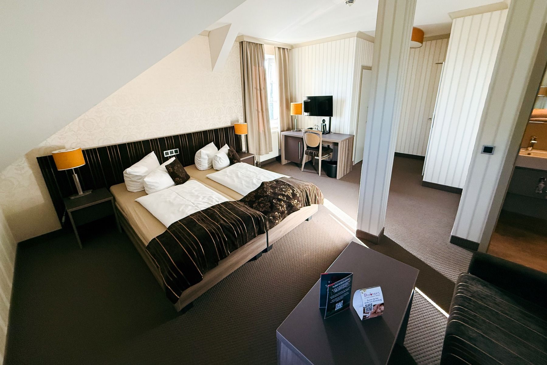 Ein Hotelzimmer im Hotel Dorotheenhof mit zwei Einzelbetten, einem Schreibtisch mit Stuhl und Fernseher, einem Sofa und einem kleinen Tisch. Warme Beleuchtung und neutrale Farben schaffen eine gemütliche, moderne Atmosphäre – perfekt zum Entspannen, nachd