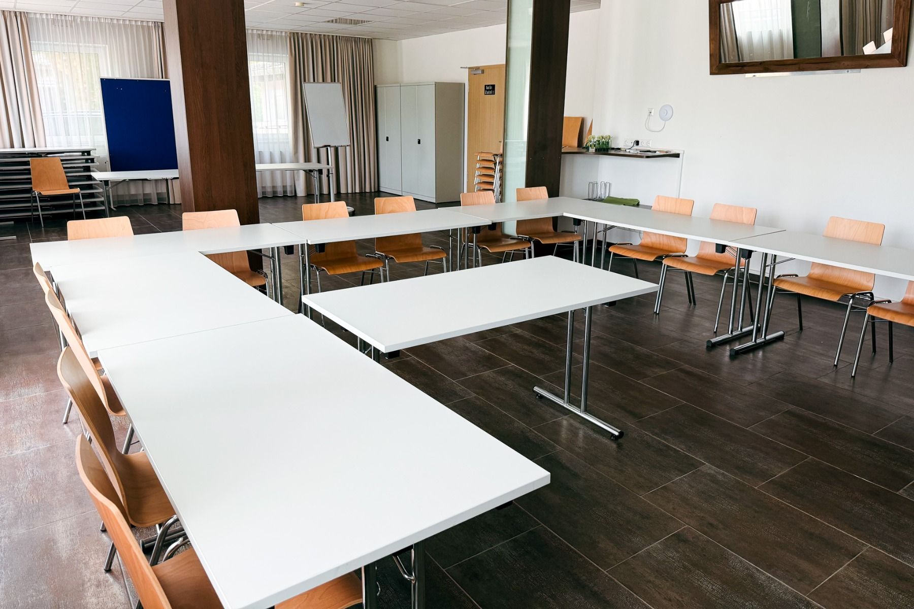 Ein heller Konferenzraum im Hotel Drei Rosen mit weißen, U-förmig angeordneten Tischen, Holzstühlen, einer blauen Präsentationswand und großen Fenstern bietet ein übersichtliches Ambiente – ideal für Fußballtrainingslager in der Nähe des Sportparks Moosbu