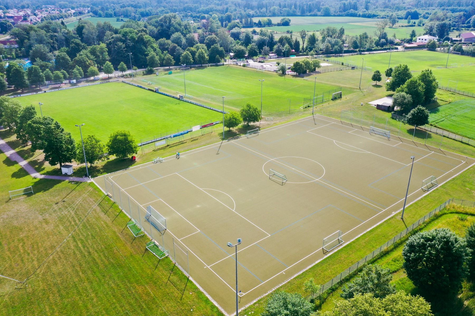 Luftaufnahme eines leeren Fußballplatzes mit Kunstrasen im Sportpark Moosburg mit Torpfosten, üppigem Rasen, Bäumen, ländlichen Gebäuden und strahlend blauem Himmel – ideal für ein Fußballtrainingslager in der Nähe des Hotels Drei Rosen.