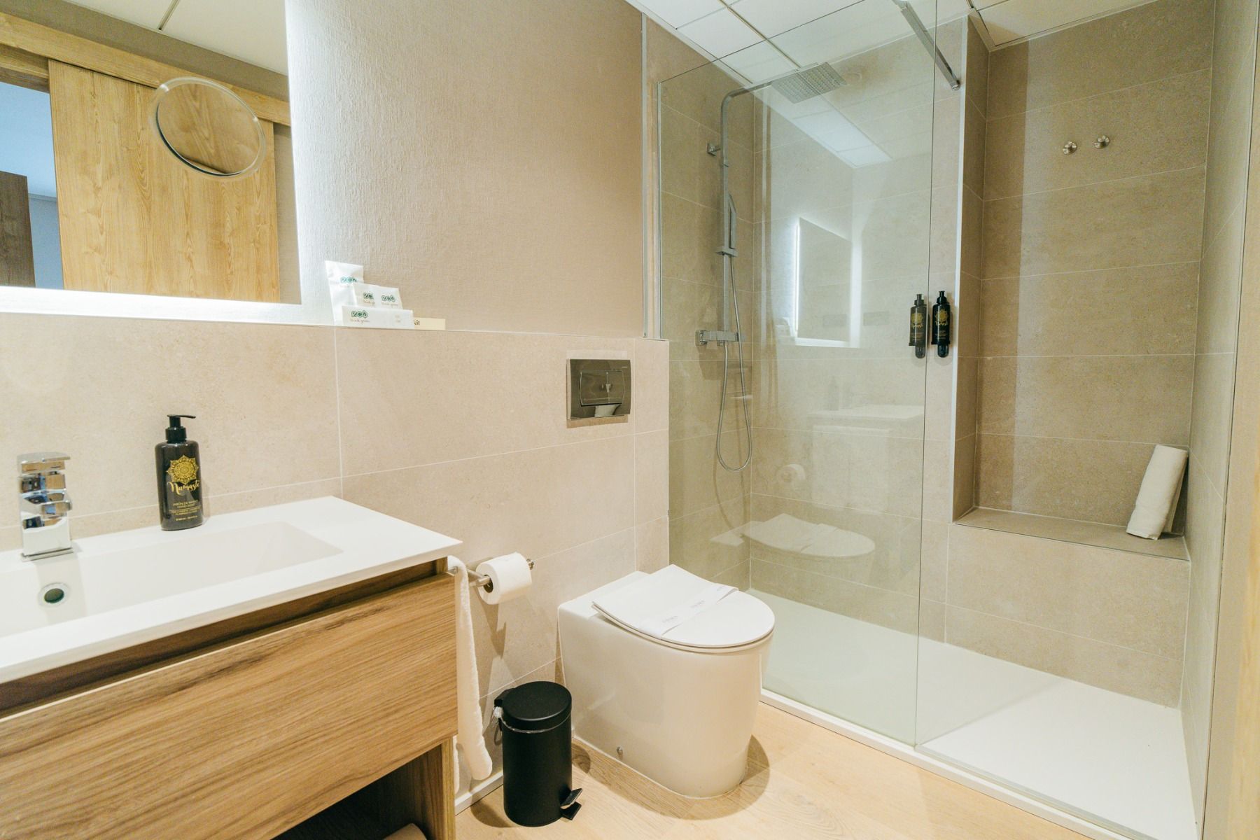 Das Hotel Gaudea bietet ein modernes Badezimmer mit einer begehbaren Dusche mit Glasabdeckung, einer Wandtoilette, einem hölzernen Waschtisch, einem Spiegel und Toilettenartikeln. Beigefarbene Fliesen und ein minimalistisches Dekor sorgen für einen entspa
