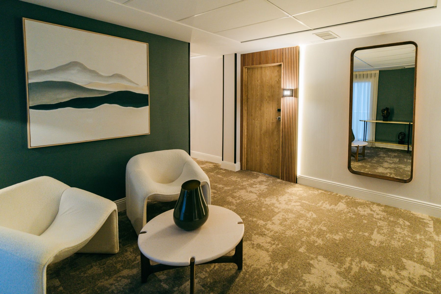 Moderne Sitzecke in neutralen Tönen im Hotel Gaudea mit zwei weißen Stühlen, einem runden Tisch mit einer dunklen Vase, großer Wandkunst, einer Holztür und einem hohen Spiegel, der das stilvolle Ambiente des Vier-Sterne-Hotels widerspiegelt.