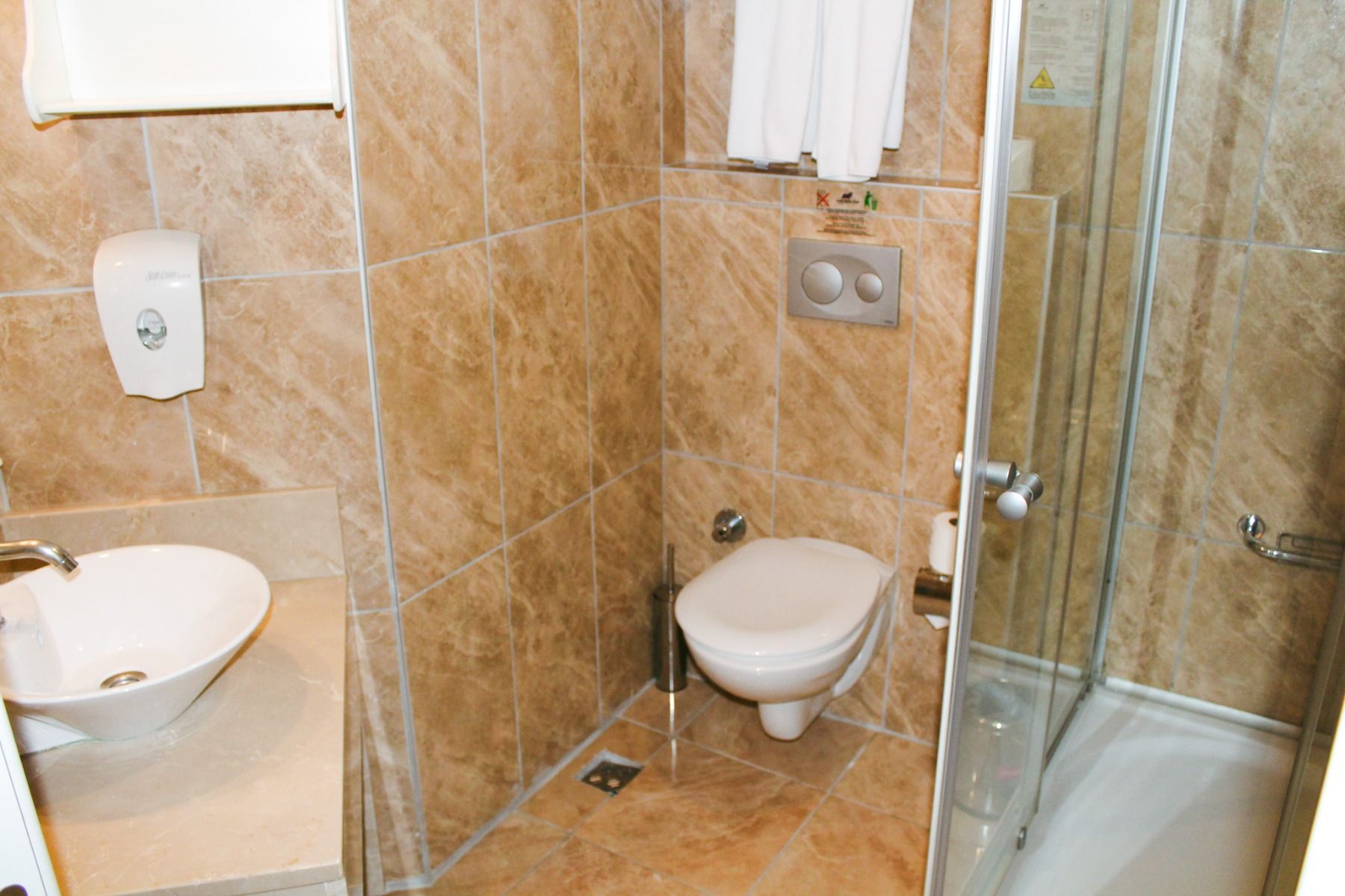 Das Badezimmer im Linda Resort ist mit hellbraunen Fliesen, einem wandmontierten WC, einer Duschkabine aus Glas, einem weißen Waschbecken mit Wasserhahn, einem Seifenspender und einem über dem WC hängenden Handtuch ausgestattet, was ihm einen modernen Loo