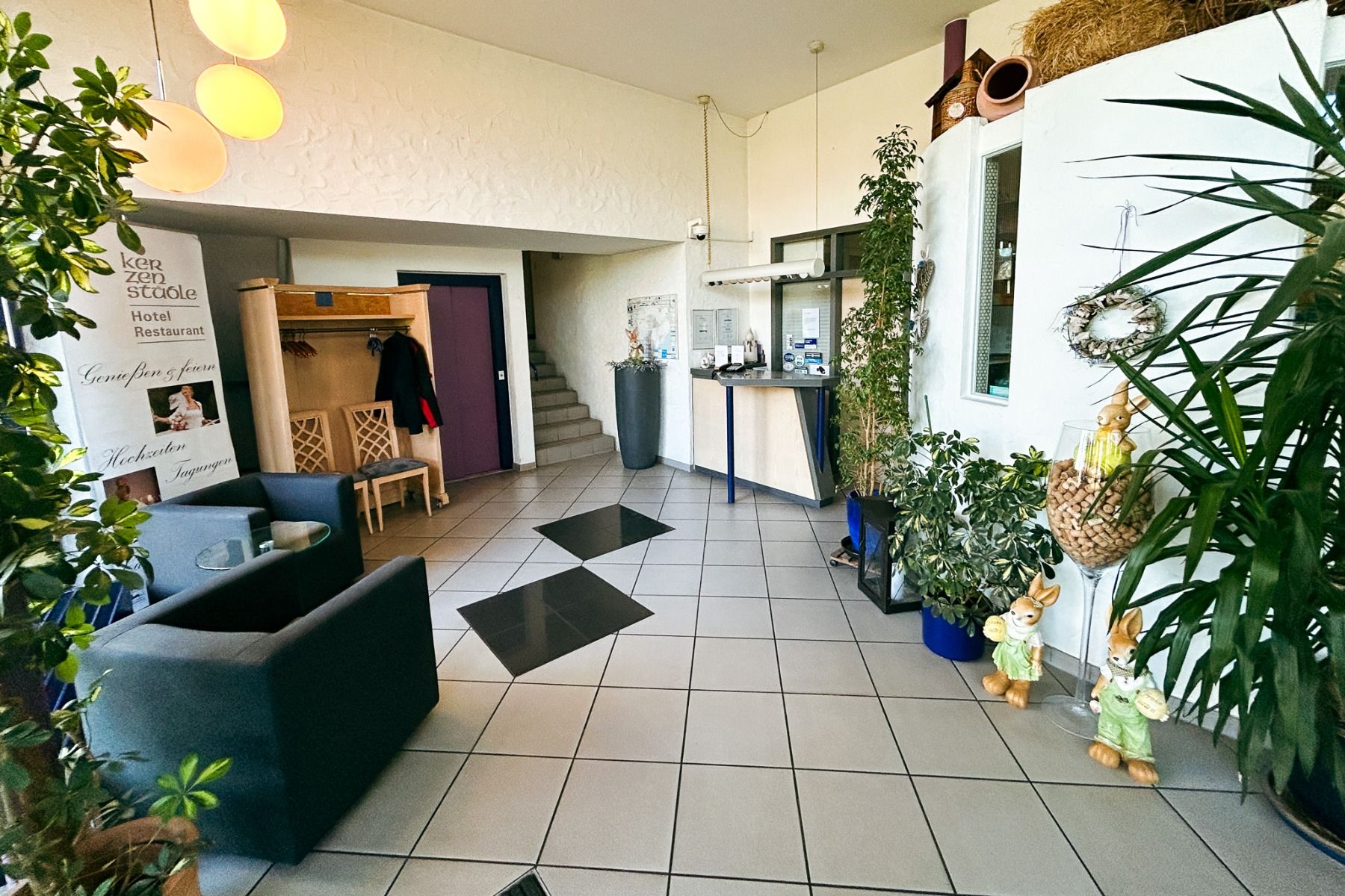 Eine helle Hotellobby heißt die Gäste im Hotel & Restaurant Kerzenstüble willkommen – mit Sesseln, üppigen Pflanzen, einer Rezeption und dekorativen Hasenfiguren. Im Hintergrund führt eine Treppe nach oben, während weiße Wände und Fliesenböden das luftige