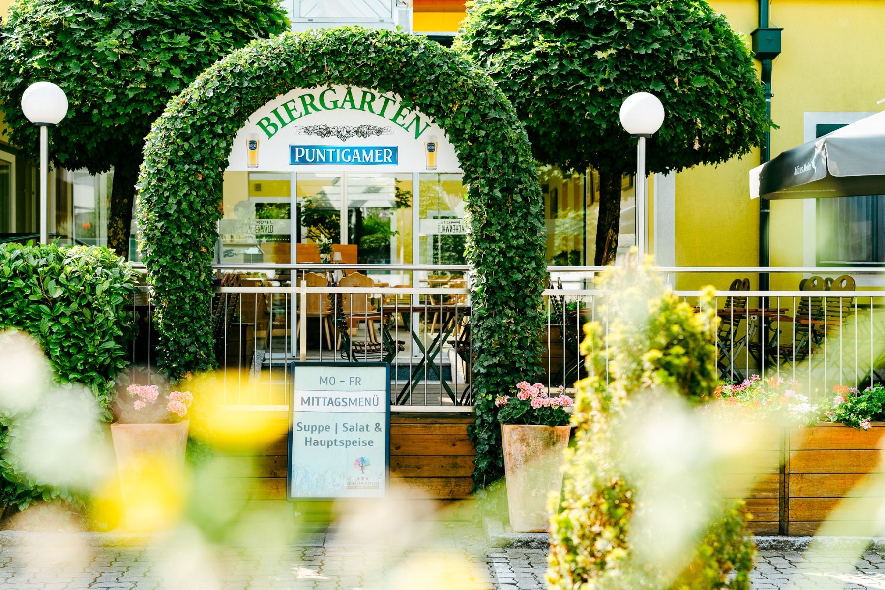 Der Eingang zum Hotel-Restaurant Schachenwald besticht durch einen grünen, begrünten Torbogen, ein Schild und eine durch Glastüren einsehbare Terrassenbereich. Zwischen Pflanzen und Blumen steht eine Speisekarte – ideal nach einem Tag am Schwarzlsee.