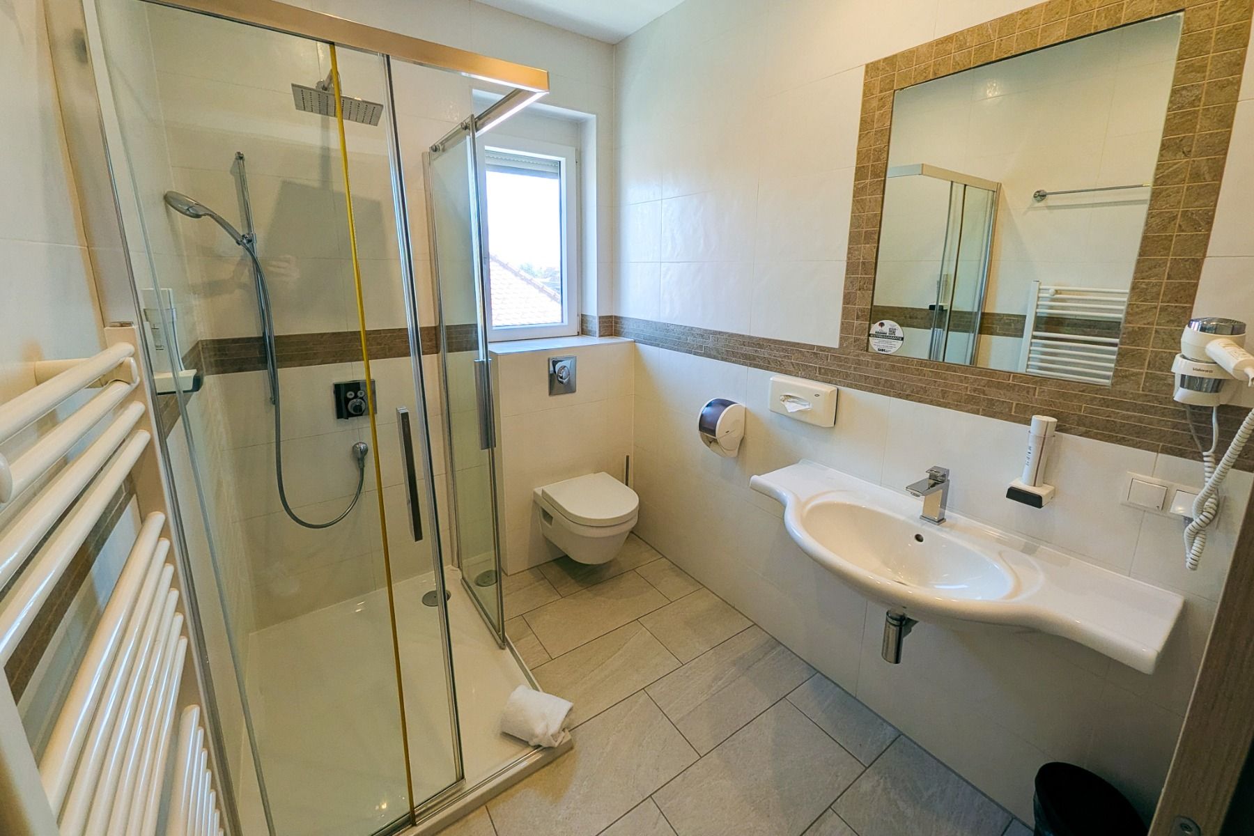 Modernes Badezimmer im Hotel Restaurant Schachenwald mit Glasduschkabine, wandhängendem WC, großem Spiegel über dem Waschbecken, Handtuchwärmer, Föhn und einem Fenster, das für Tageslicht sorgt – ideal zum Entspannen nach einem Tag am Schwarzlsee.