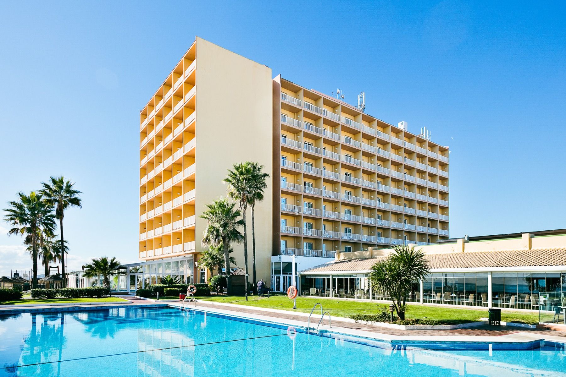 Ein hohes, gelbes Hotel Sol Guadalmar mit Balkonen blickt auf einen klaren, blauen Außenpool, umgeben von Palmen und grünen Rasenflächen unter einem sonnigen Himmel - perfekt für Ihren Aufenthalt in diesem zentrumsnahen 4 Sterne Hotel Malaga.