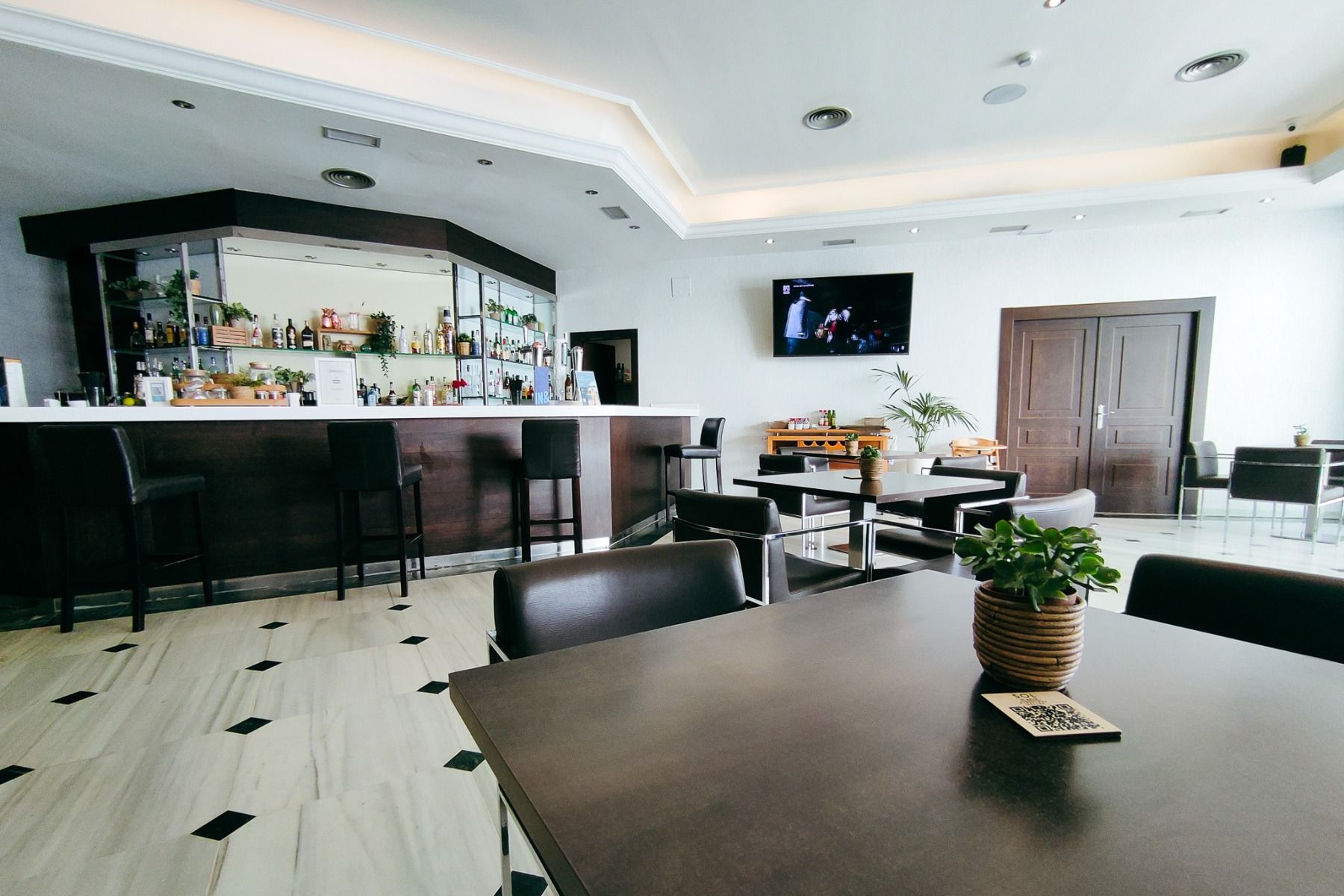Die moderne Bar im Hotel Sol Guadalmar in Málaga besteht aus einem weißen Tresen, schwarzen Stühlen und Tischen. Eine kleine Pflanze schmückt den nächsten Tisch. Hinter der Bar stehen Flaschen und an der weißen Wand ist ein Fernseher angebracht.