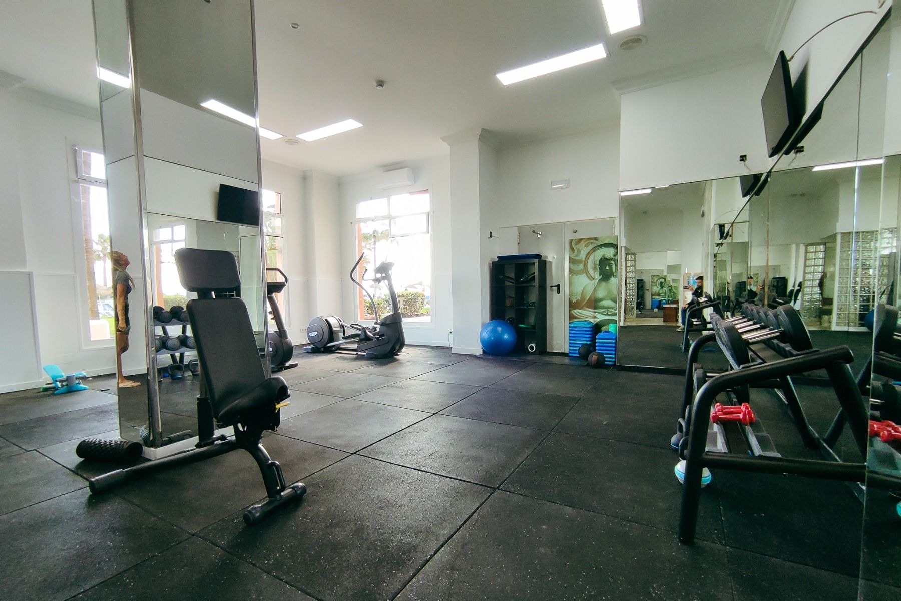 Der Fitnessraum im Hotel Sol Guadalmar ist mit Spiegeln, Heimtrainern, einer Hantelbank, freien Gewichten, Gymnastikbällen und Matten in Regalen ausgestattet. Große Fenster erhellen den Raum mit natürlichem Licht.