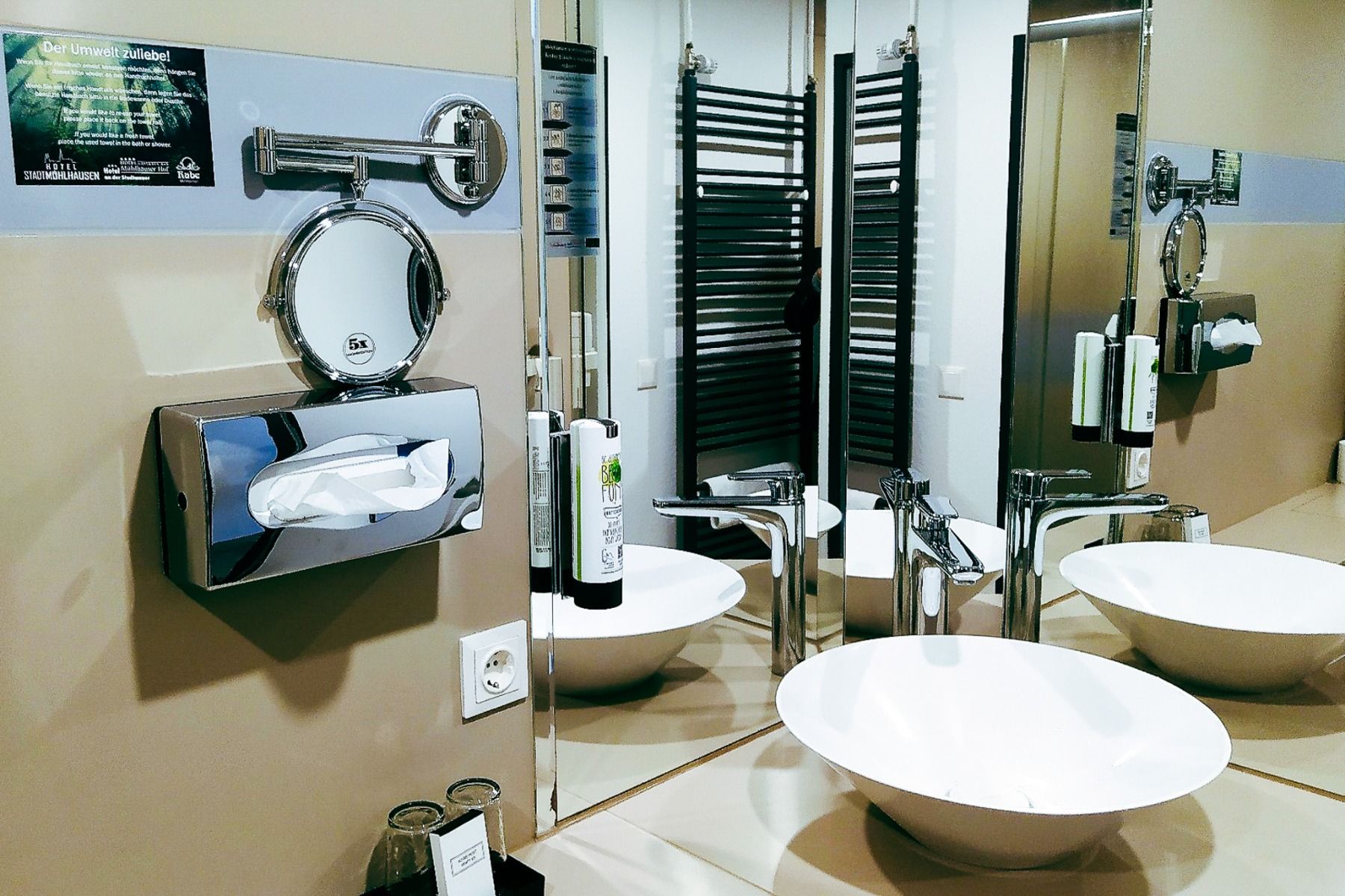 Ein modernes Badezimmer mit einem weißen Aufsatzwaschbecken, einer hohen Armatur, einem an der Wand montierten Toilettenpapierspender und einem runden Vergrößerungsspiegel – stilvolle Ausstattungsmerkmale, die Sie im Hotel Stadt Mühlhausen*** vorfinden.