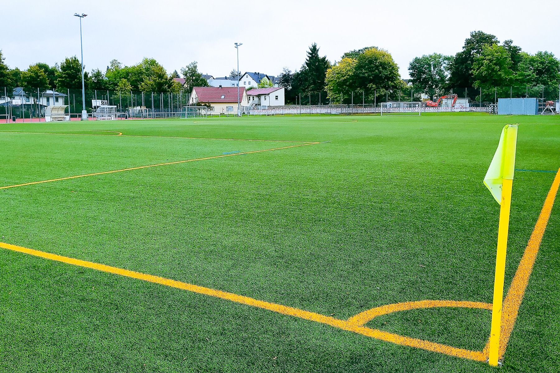 Ein hellgrüner Kunstrasen-Fußballplatz mit gelben Linien und einer Eckfahne liegt in der Nähe des Hotels Stadt Mühlhausen***, umgeben von Bäumen, Häusern und Sportanlagen – darunter ein Fitnessstudio – unter bewölktem Himmel.