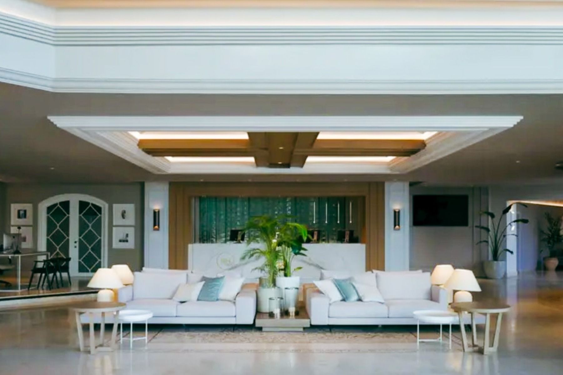 Die moderne Lobby des Hotels SH Villa Gadea* ist mit weißen Sofas, Beistelltischen mit Lampen, einer großen Zimmerpflanze in der Mitte und einer Rezeption unter einer Kassettendecke ausgestattet – ideal für Fußballmannschaften auf Trainingslager in Altea.