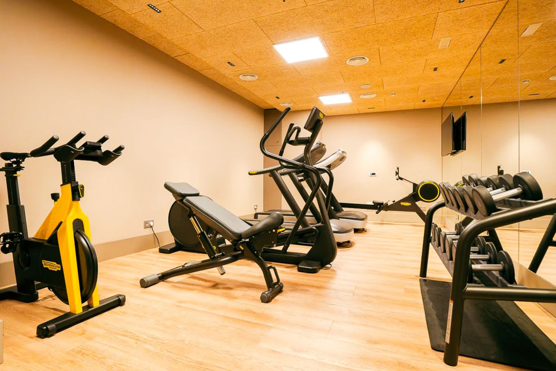 Das Hotel Zenit Sevilla verfügt über ein modernes Fitnessstudio mit stationären Fahrrädern, Bänken, elliptischen Maschinen, Hanteln auf einer Ablage, Holzfußböden, großen Spiegeln und hellen Deckenleuchten - ideal für Fußball-Sportgruppen, um fit zu bleib