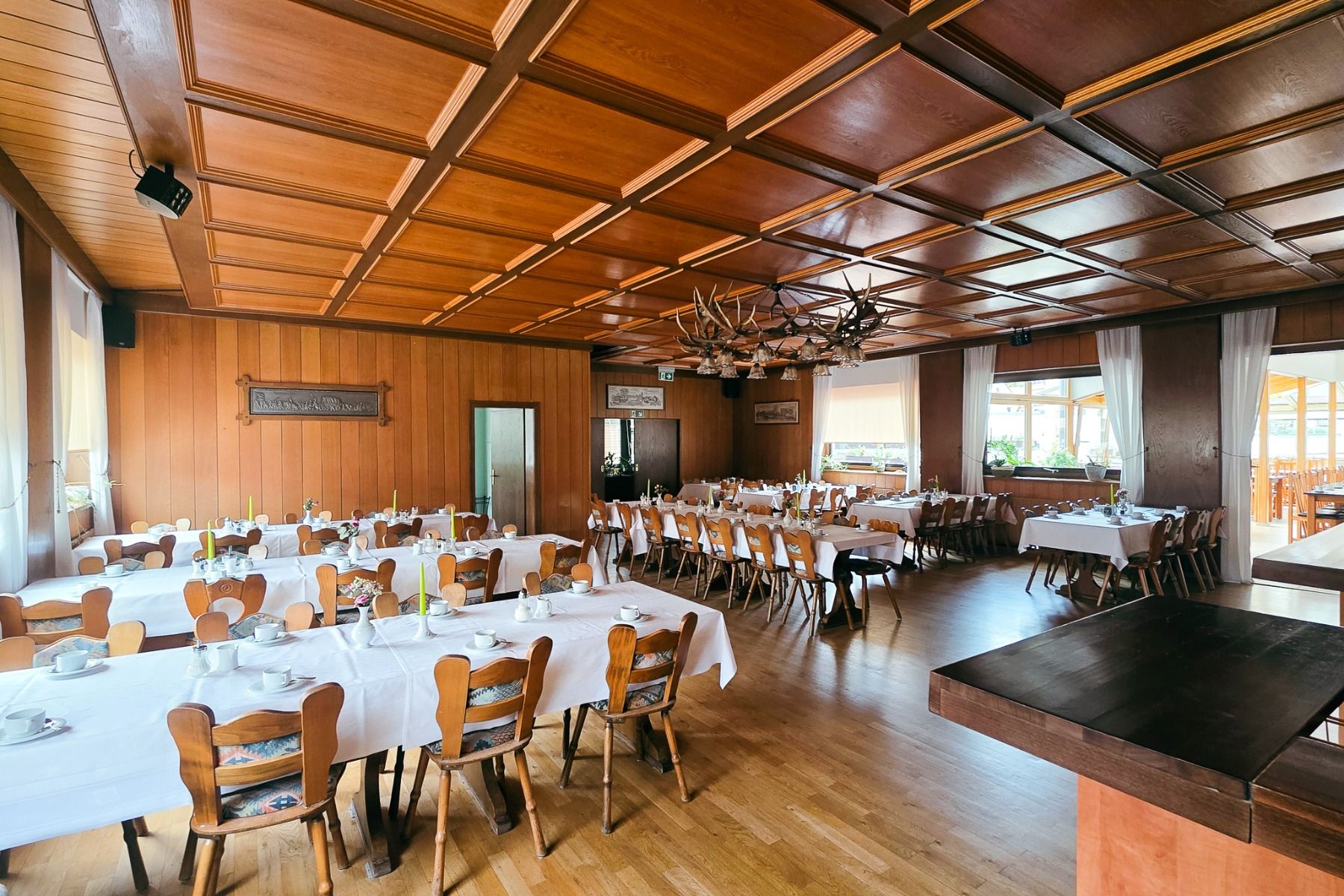 Der geräumige, holzgetäfelte Speisesaal des Gasthofs Kapellenhof verfügt über ordentlich gedeckte Tische mit weißen Tüchern, Holzstühle, große, sonnendurchflutete Fenster und einen dekorativen Kronleuchter - ideal, um ein gemütliches Ambiente zu schaffen.
