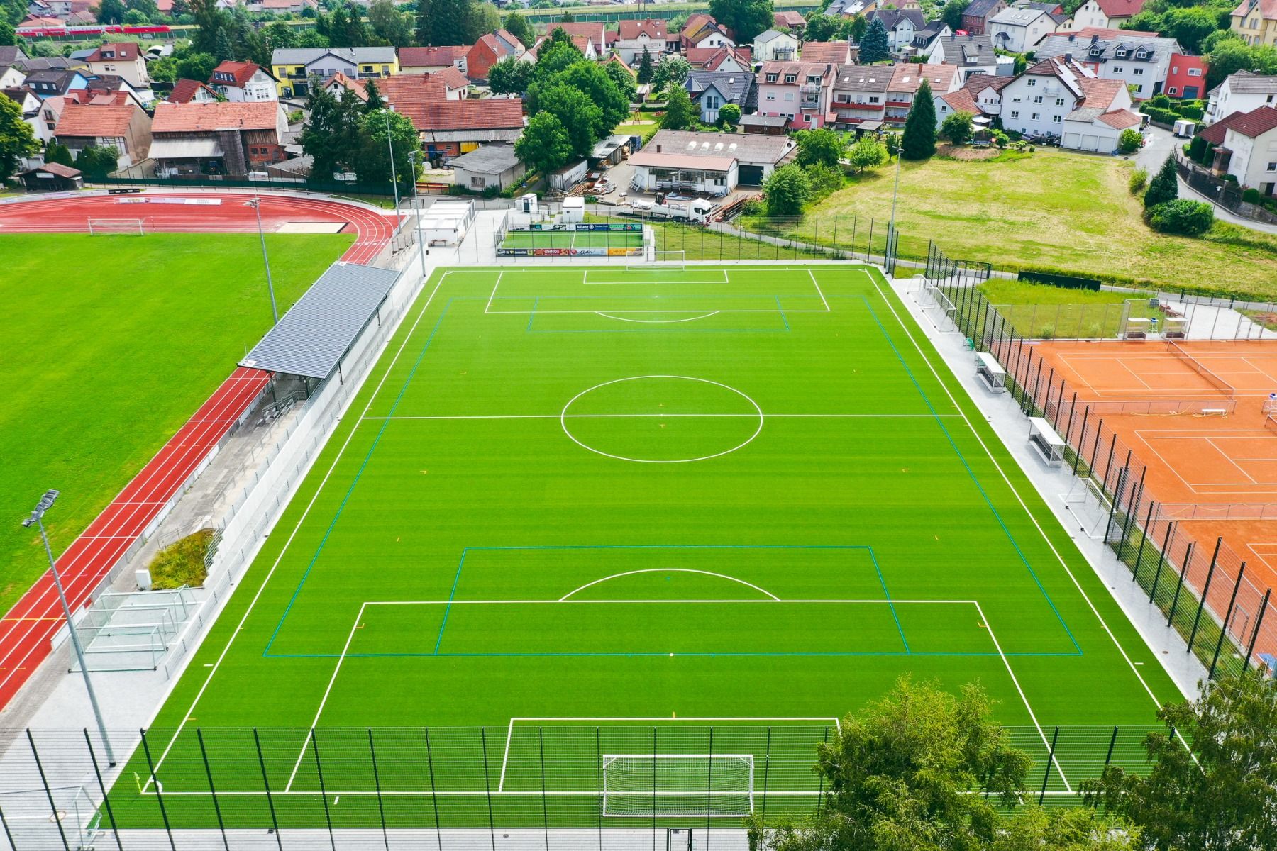 Luftaufnahme eines grünen Fußballplatzes – ideal für ein Trainingslager – mit weißen und blauen Markierungen, neben einer Laufbahn und orangefarbenen Tennisplätzen in der Nähe des Landhotels Imhof in einer ruhigen Wohngegend mit Häusern und Bäumen.