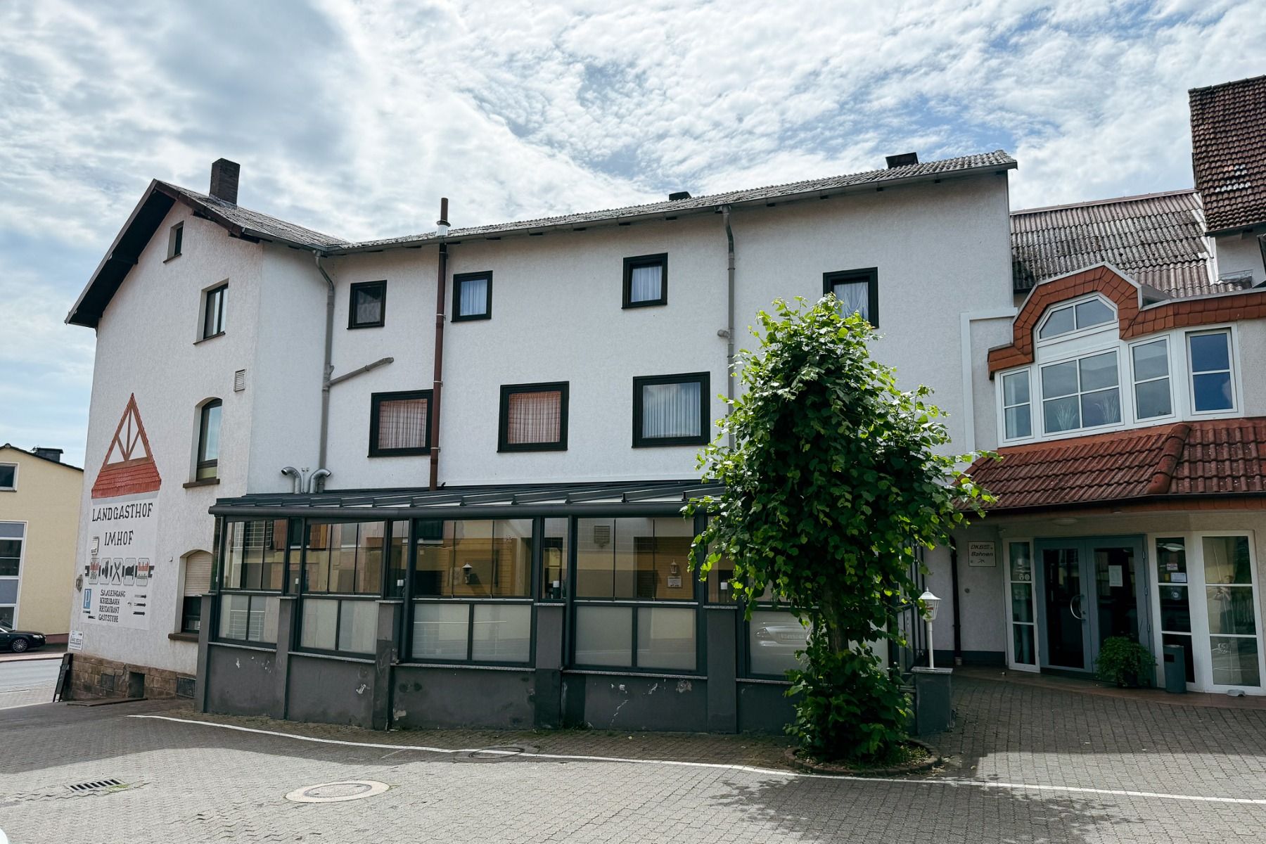 Ein weißes, dreistöckiges Gebäude mit braunen Dachziegeln, vielen Fenstern, einer verglasten Terrasse und einem üppig belaubten Baum davor. Auf der linken Seite steht auf einem Schild „Landhotel Imhof“. Dieses familiengeführte Hotel eignet sich ideal für 