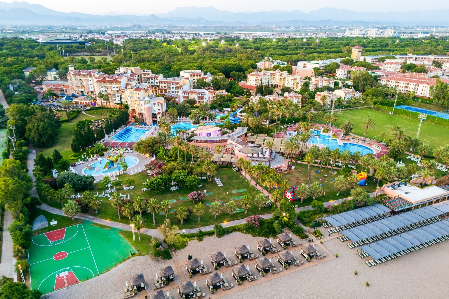 Luftaufnahme des Hotels Limak Arcadia mit Pools, Gärten, Sportplätzen und Strandkabinen - ideal für das Fußballtrainingslager Türkei - inmitten von Palmen und Bergen unter einem klaren Himmel.