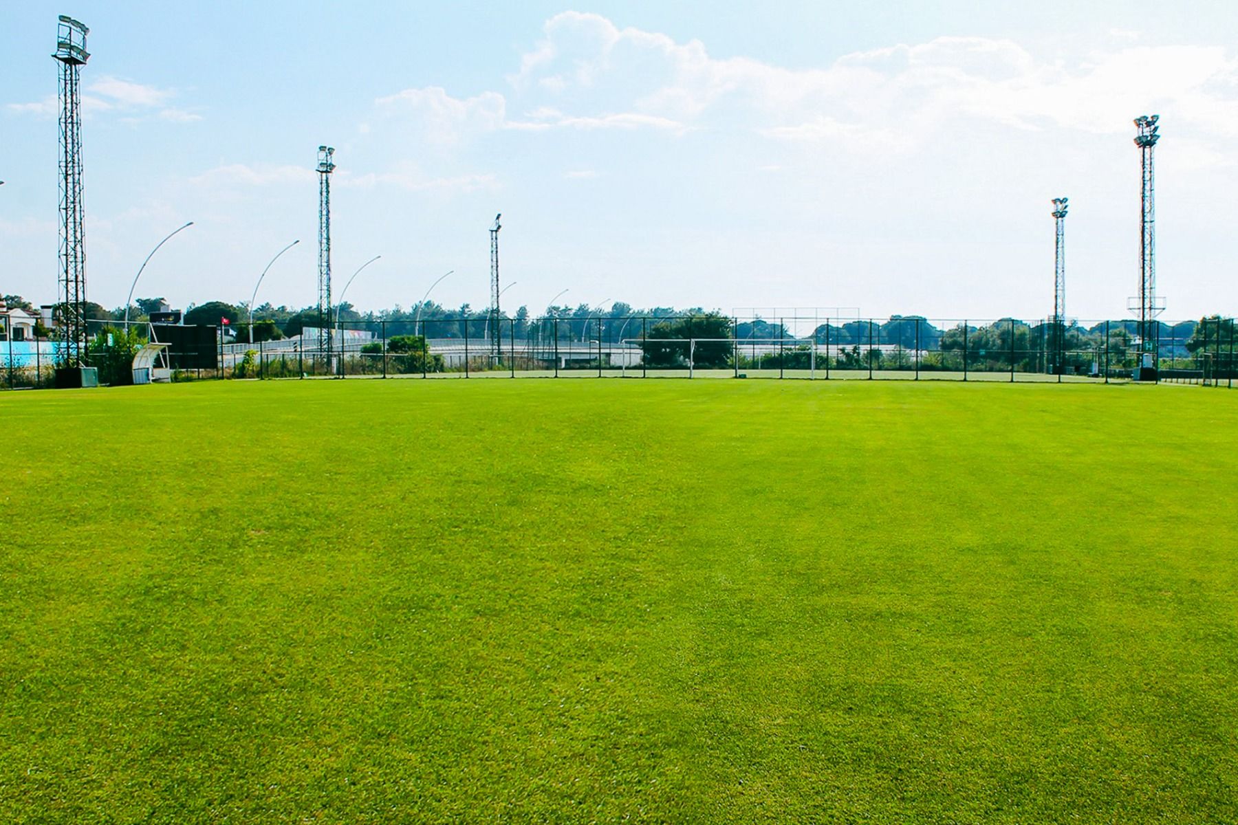 Ein großer, gepflegter grüner Sportplatz am Hotel Limak Arcadia unter strahlendem Himmel - perfekt für ein Fußballtrainingslager Türkei- mit Flutlicht, Metallzäunen und Blick auf Bäume und Gebäude im Hintergrund.