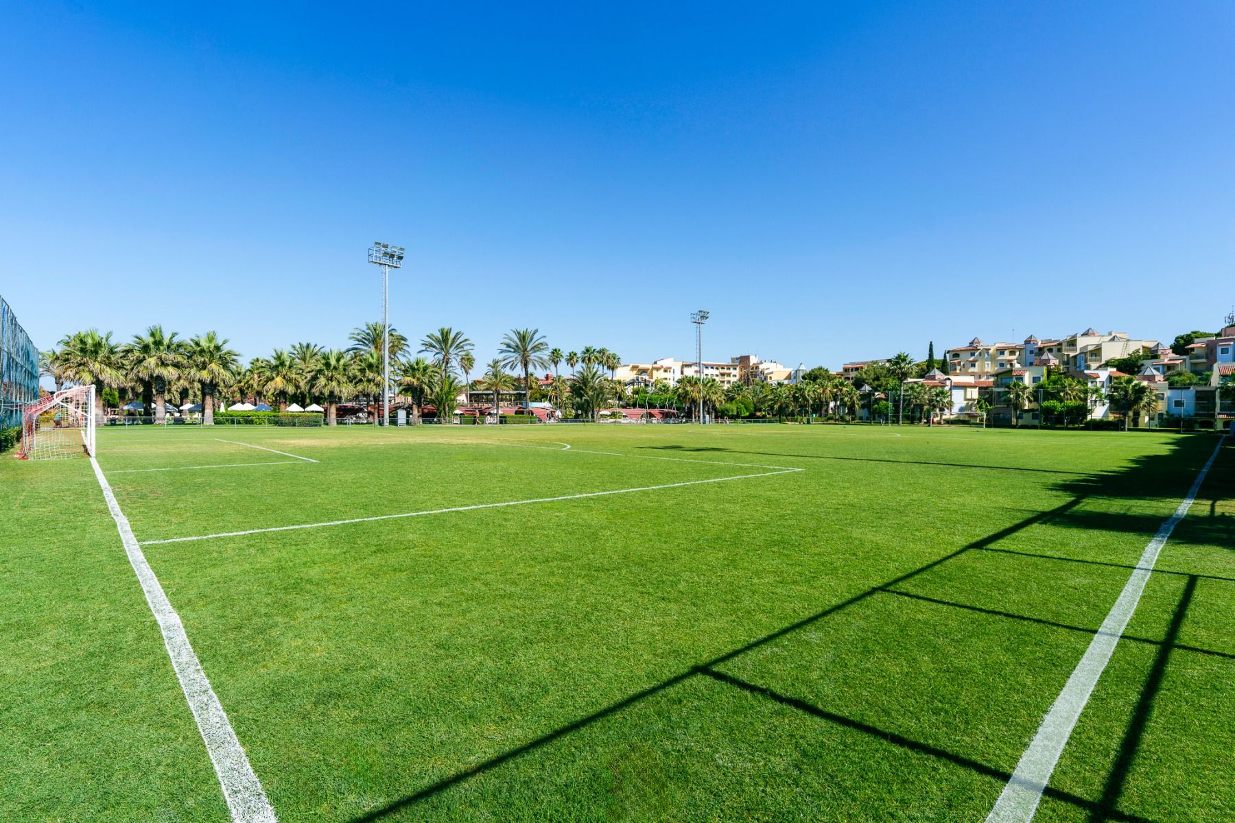 Ein sonniger Fußballplatz im Freien beim Hotel Limak Arcadia in der Türkei, ideal für ein Fußballtrainingslager, mit grünem Rasen, weißen Linien, einem linken Torpfosten, Palmen und Gebäuden unter einem klaren blauen Himmel.