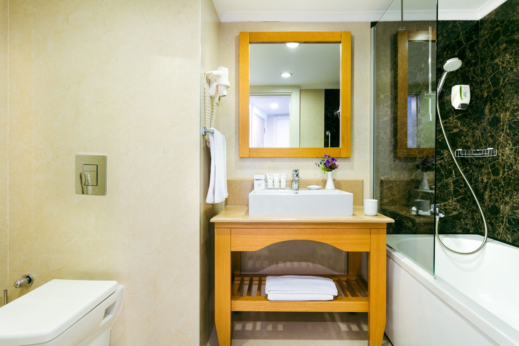 Das moderne Badezimmer im Hotel Limak Arcadia verfügt über einen hölzernen Waschtisch, ein weißes Waschbecken, einen Spiegel, Toilettenartikel, eine Toilette auf der linken Seite, eine Badewanne mit Dusche auf der rechten Seite und eine Glasabtrennung, di