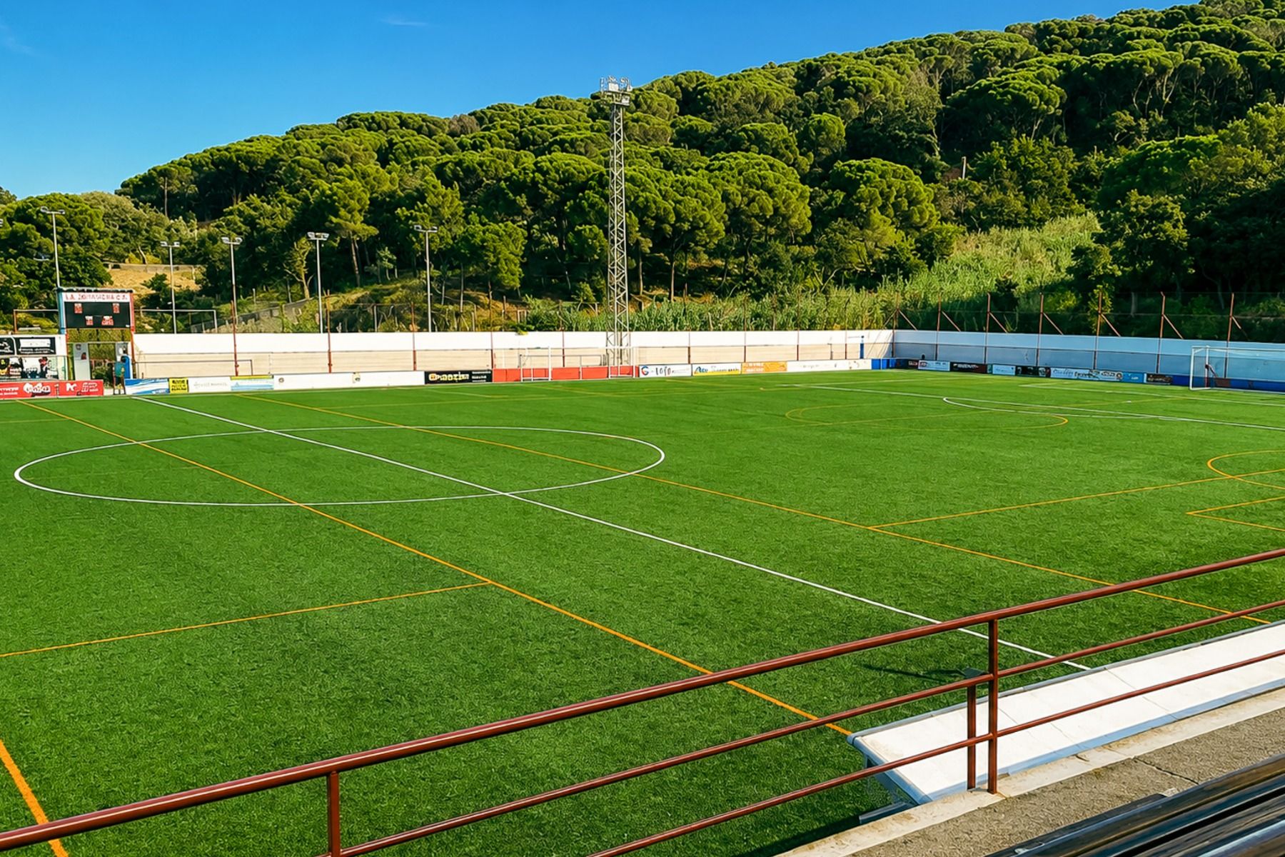 Am Van der Valk Hotel BARCAROLA in Costa Brava gibt es einen leeren Fußballplatz mit grünem Kunstrasen, klaren Markierungen und Sitzgelegenheiten - perfekt für ein Fußball-Trainingslager zwischen Bäumen, Hügeln und blauem Himmel.