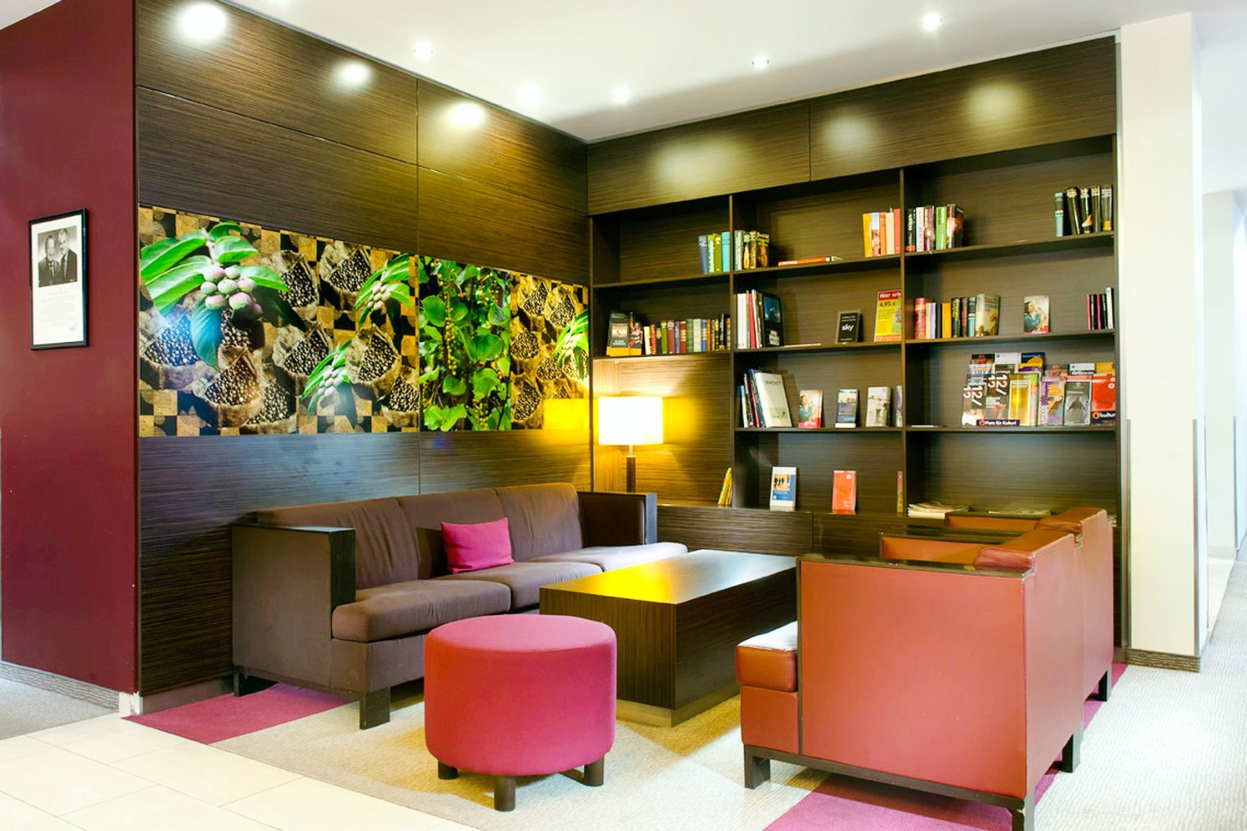 Die moderne Lounge im Mercure Hotel ist mit braunen Sofas, roten Stühlen, einem Couchtisch aus Holz und einem Bücherregal voller Bücher und Zeitschriften ausgestattet – ideal, um sich nach einem Tag in Ihrem Trainingslager zu entspannen. Bunte Wandbilder 