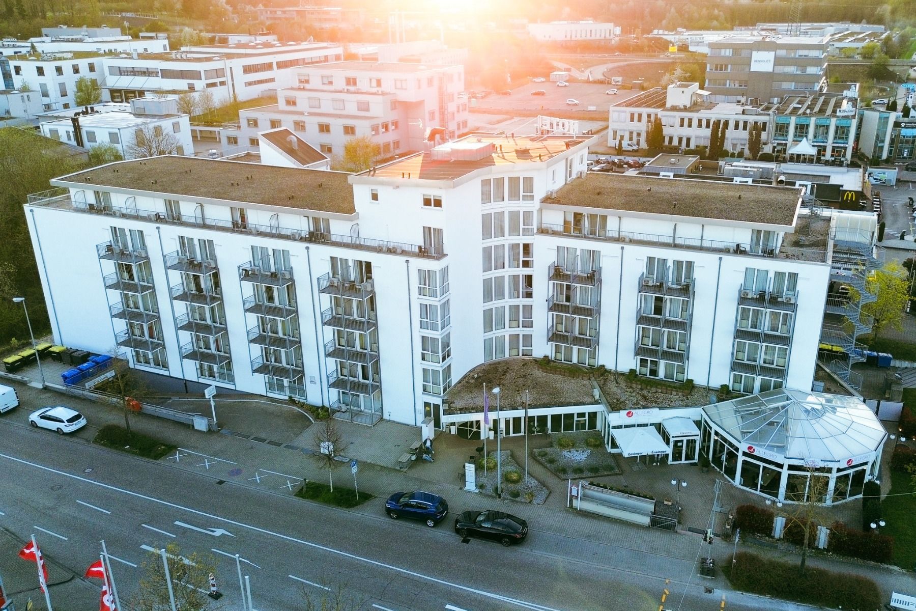 Luftaufnahme des Mercure Hotels Pforzheim, eines modernen, weißen Hotels mit Balkonen, einem gläsernen Atrium und einem Parkplatz vor dem Haus, eingebettet zwischen den Gebäuden der Stadt und im Sonnenlicht – dank seiner guten Erreichbarkeit ideal für Bes