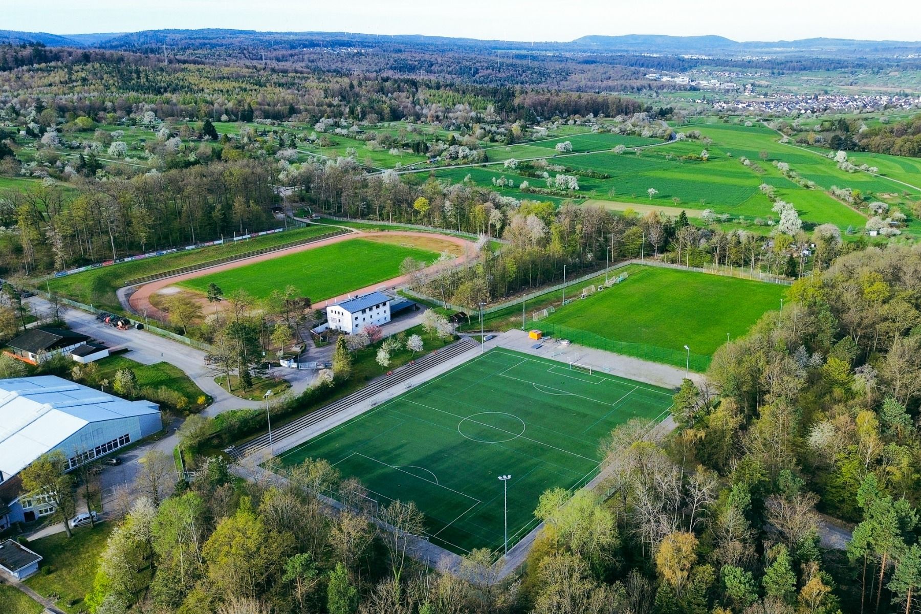 Luftaufnahme eines Sportkomplexes mit Kunstrasenplätzen, einer Laufbahn und umliegenden Bäumen, eingebettet in eine ländliche, grüne Landschaft mit Hügeln und vereinzelten Gebäuden – ideal für ein Trainingslager in Pforzheim aufgrund der guten Erreichbark