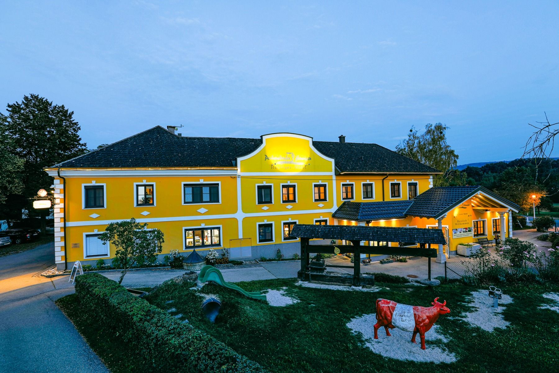Das Perbersdorfer Heuriger - Hotel in Neuhofen an der Ybbs zeigt sich in der Abenddämmerung mit seiner leuchtend gelben Fassade und den weißen Verkleidungen in warmem Licht. Eine rot-weiße Kuhskulptur ruht auf Kies inmitten von Grün in der Nähe des einlad