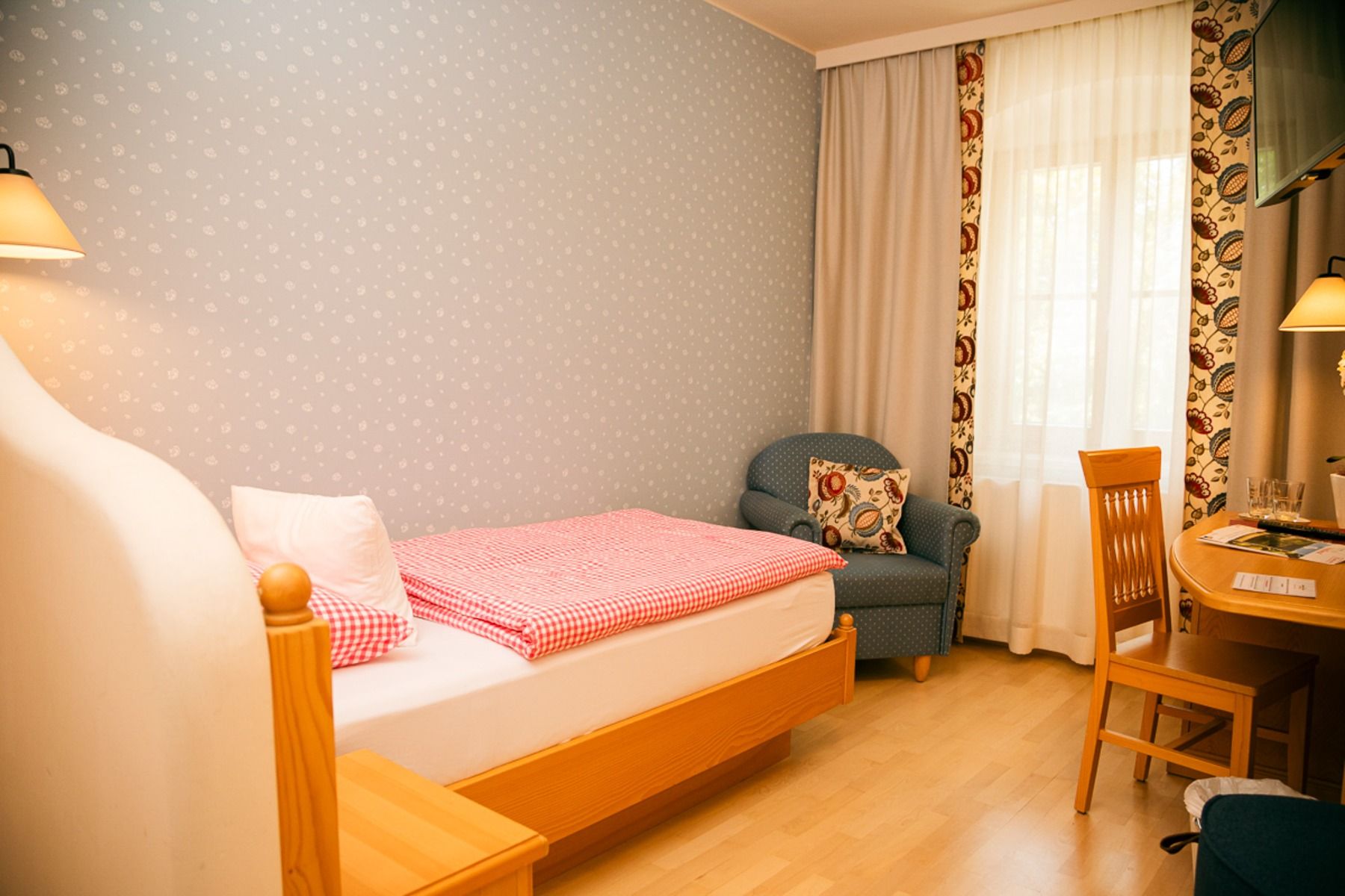 Ein gemütliches Schlafzimmer im Perbersdorfer Heurigen - Hotel mit einem Einzelbett mit rot karierter Bettwäsche, einem kleinen Holzschreibtisch und -stuhl, einem gepolsterten Sessel, Holzböden, tapezierten Wänden und einem Fenster mit durchsichtigen Vorh
