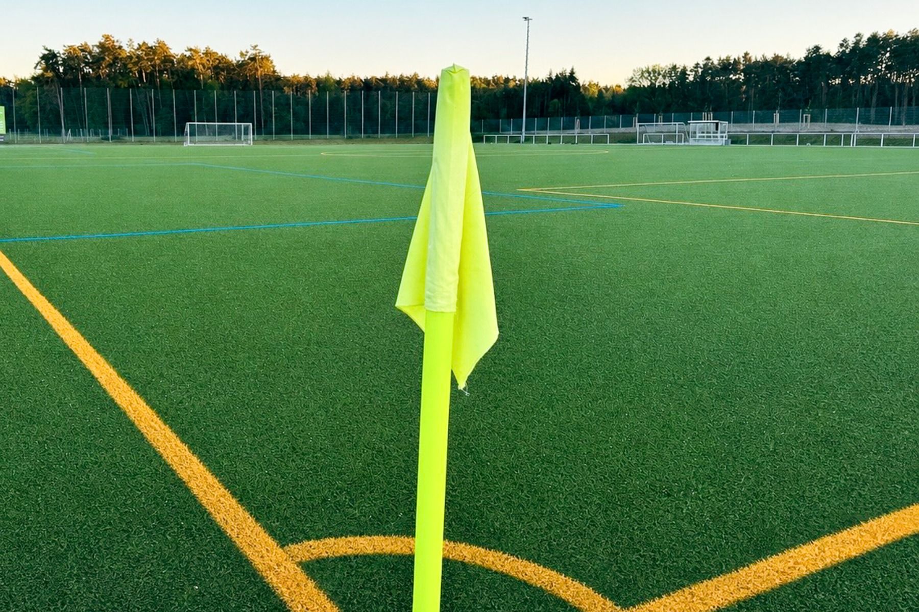 Eine gelbe Eckfahne steht am Rande eines leeren Kunstrasen-Fußballplatzes mit orangefarbenen und blauen Linien, während Torpfosten und Bäume das gemütliche Ambiente beim Gasthof Kapellenhof unter freiem Himmel verschönern.