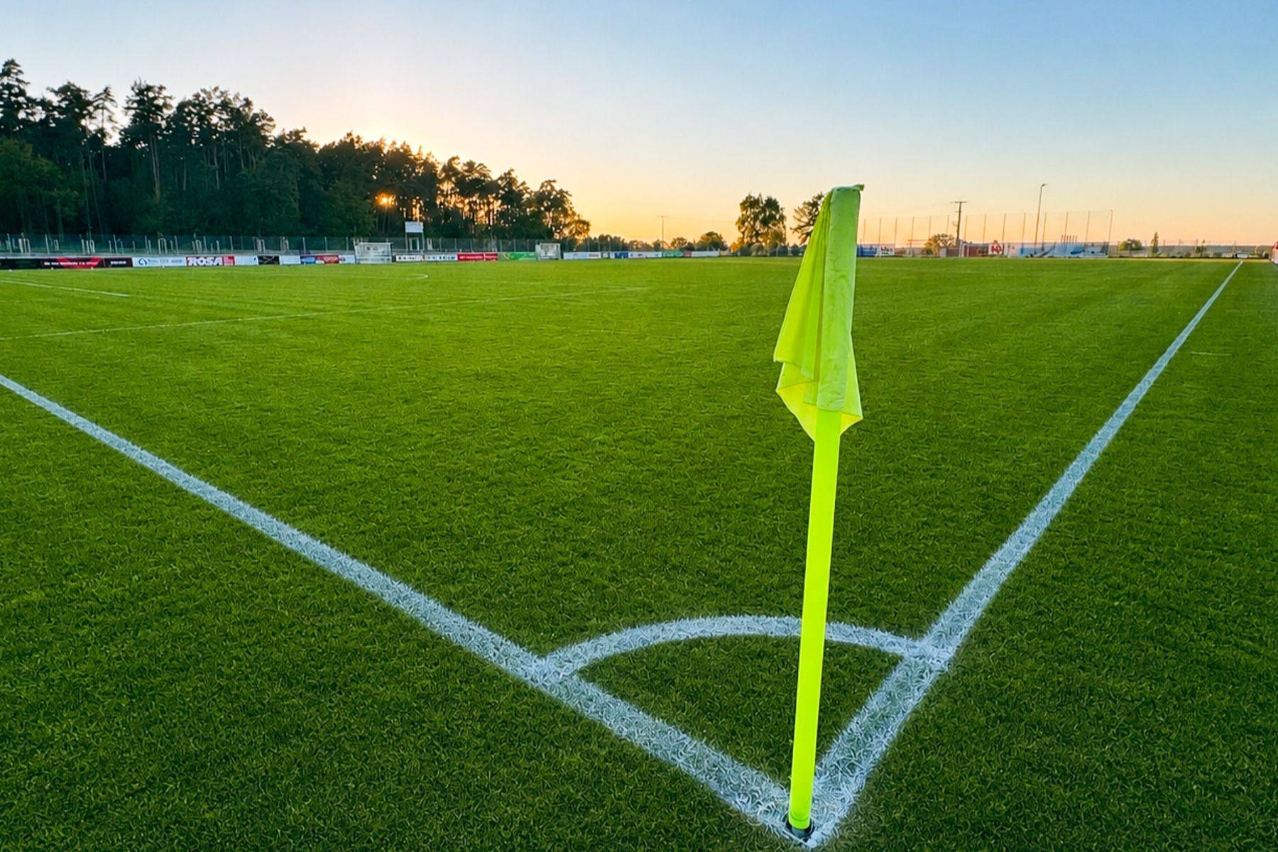 Eine leuchtend gelbe Eckfahne steht bei Sonnenuntergang auf einem gepflegten Fußballplatz in der Nähe des Gasthofs Kapellenhof, mit weißen Linien und Bäumen im Hintergrund, die ein gemütliches Ambiente unter dem blauen Himmel hervorrufen.