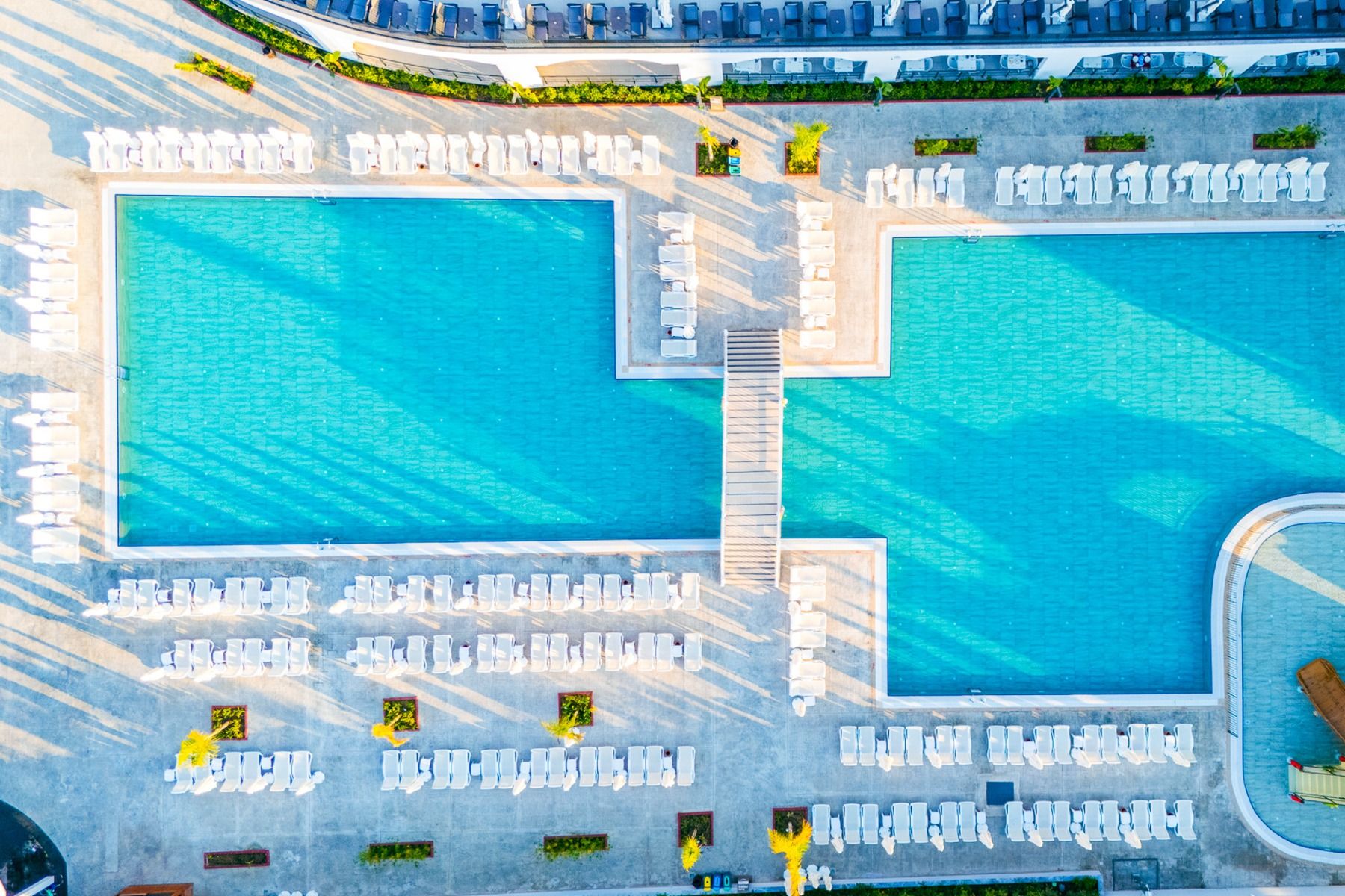 Luftaufnahme des großen, rechteckigen Pools des Side Moon Palace mit einer Verbindungsbrücke, umgeben von ordentlich aufgereihten weißen Liegestühlen auf der sonnenbeschienenen Terrasse.